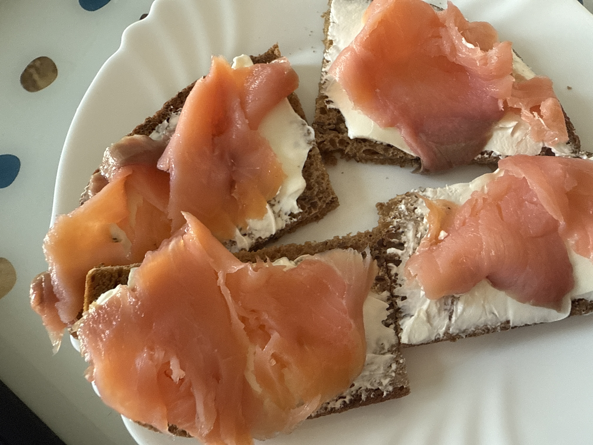 A close-up overhead shot shows four open-faced sandwiches on a white plate. The sandwiches are made with dark rye bread, a layer of cream cheese, and slices of smoked salmon. The salmon is a vibrant pink-orange color and appears thinly sliced. The plate has a scalloped edge and is on a surface with blue and brown dots. The lighting is bright, and the focus is sharp on the food.