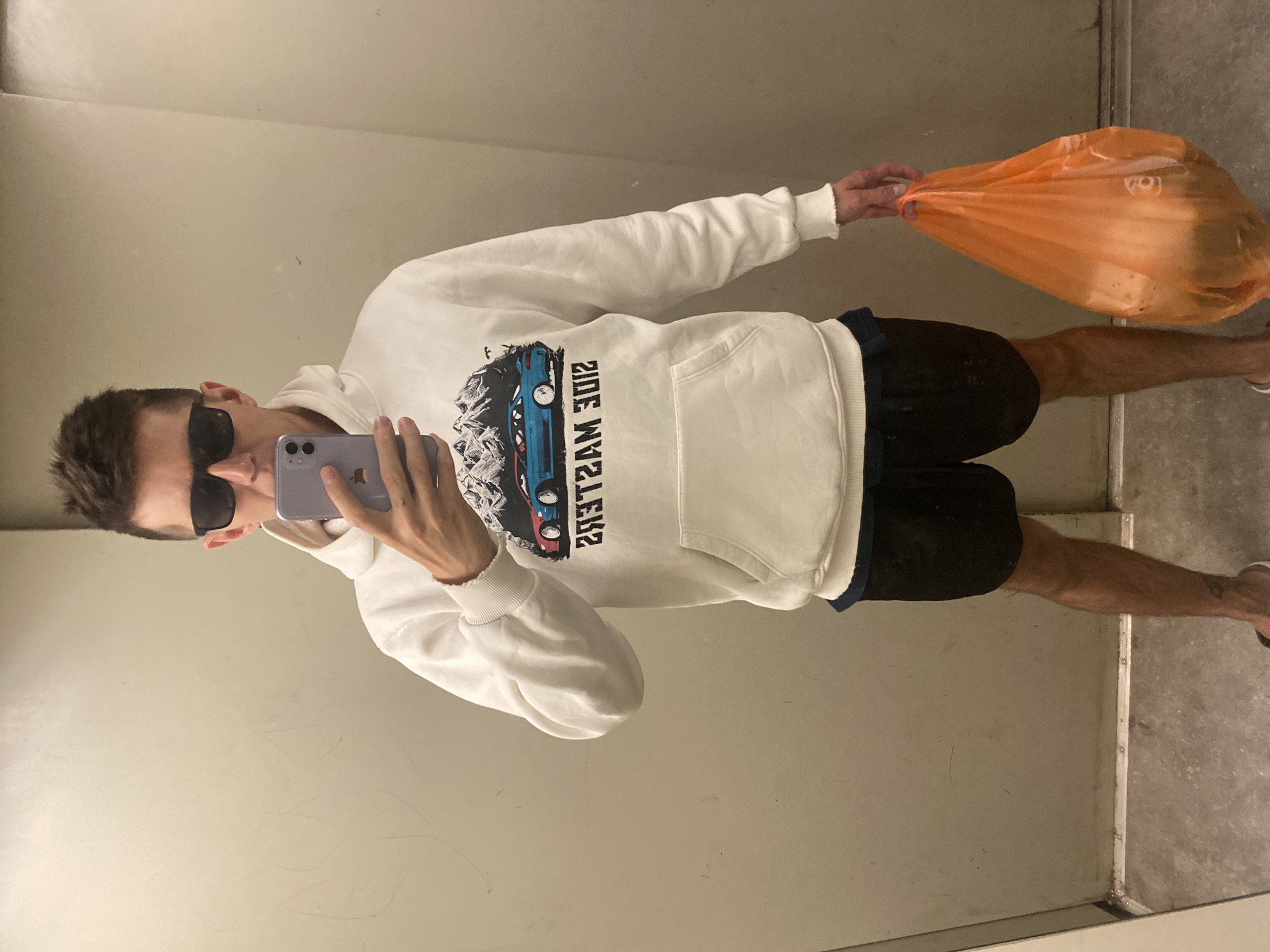 A young adult male with short, dark hair and sunglasses is standing in what appears to be a mirrored room with gray walls and a gray floor. He is wearing a white hooded sweatshirt with a graphic of two cars and mountains on the front, and black shorts. He is holding a smartphone in his right hand, taking a selfie. In his left hand, he holds a bright orange plastic bag filled with items. The text on the sweatshirt is backward, indicating it's a reflection. The overall scene suggests a casual, indoor environment, possibly a fitting room or a public restroom with mirrors. The location in Novokuybyshevsk, Russia is not discernible from the visual information provided.