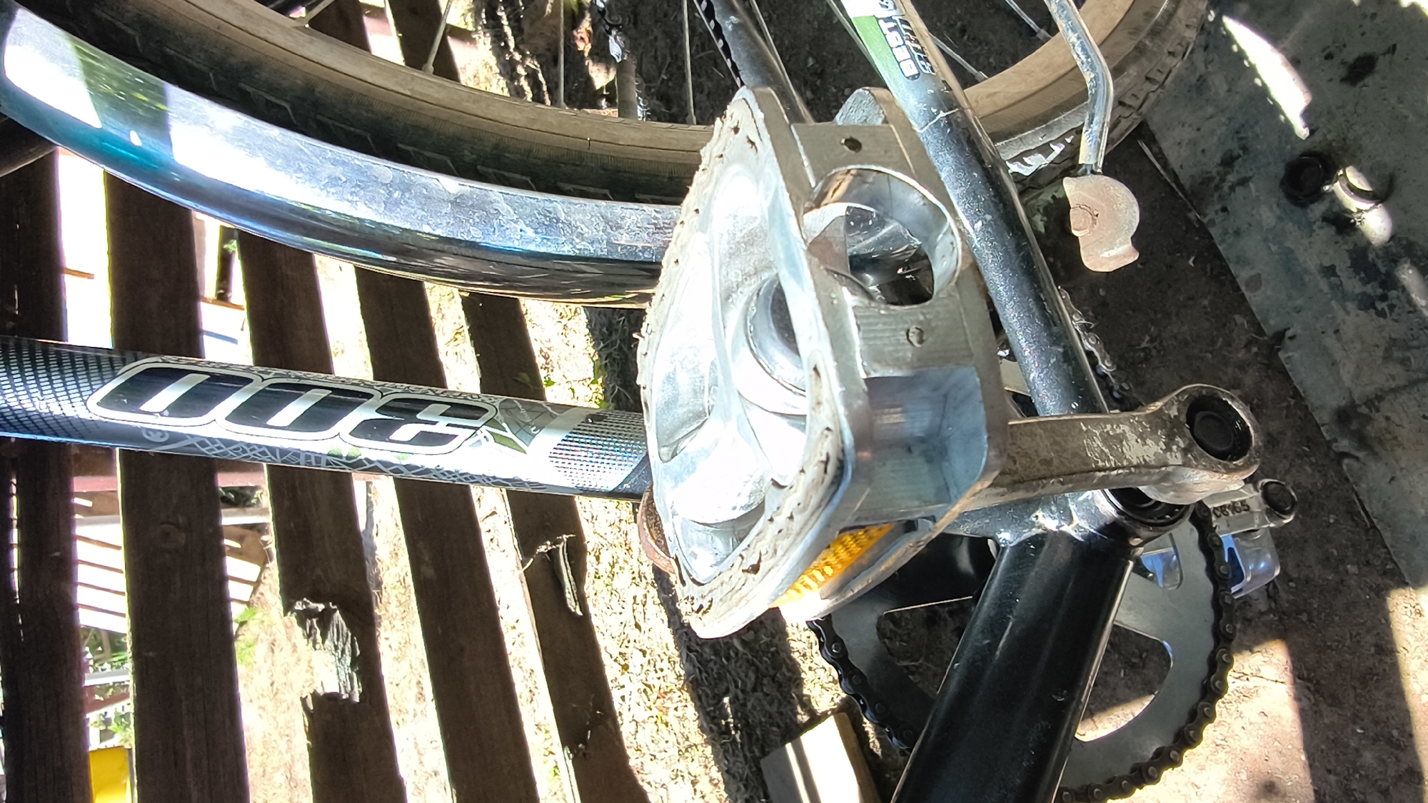 The image shows a close-up of a bicycle pedal and part of the frame, outdoors. The bicycle appears to be parked or resting against a wooden fence. The pedal is made of metal with a plastic reflector, and it is attached to a crank arm. The chainring and part of the chain are visible. The bicycle's frame is black, and a portion of the rear wheel and fender can be seen. The ground is covered with dirt and debris. The sunlight suggests it is daytime. The image does not contain any visual cues that would place it specifically in the city of Mikhaylovka, Russia.
