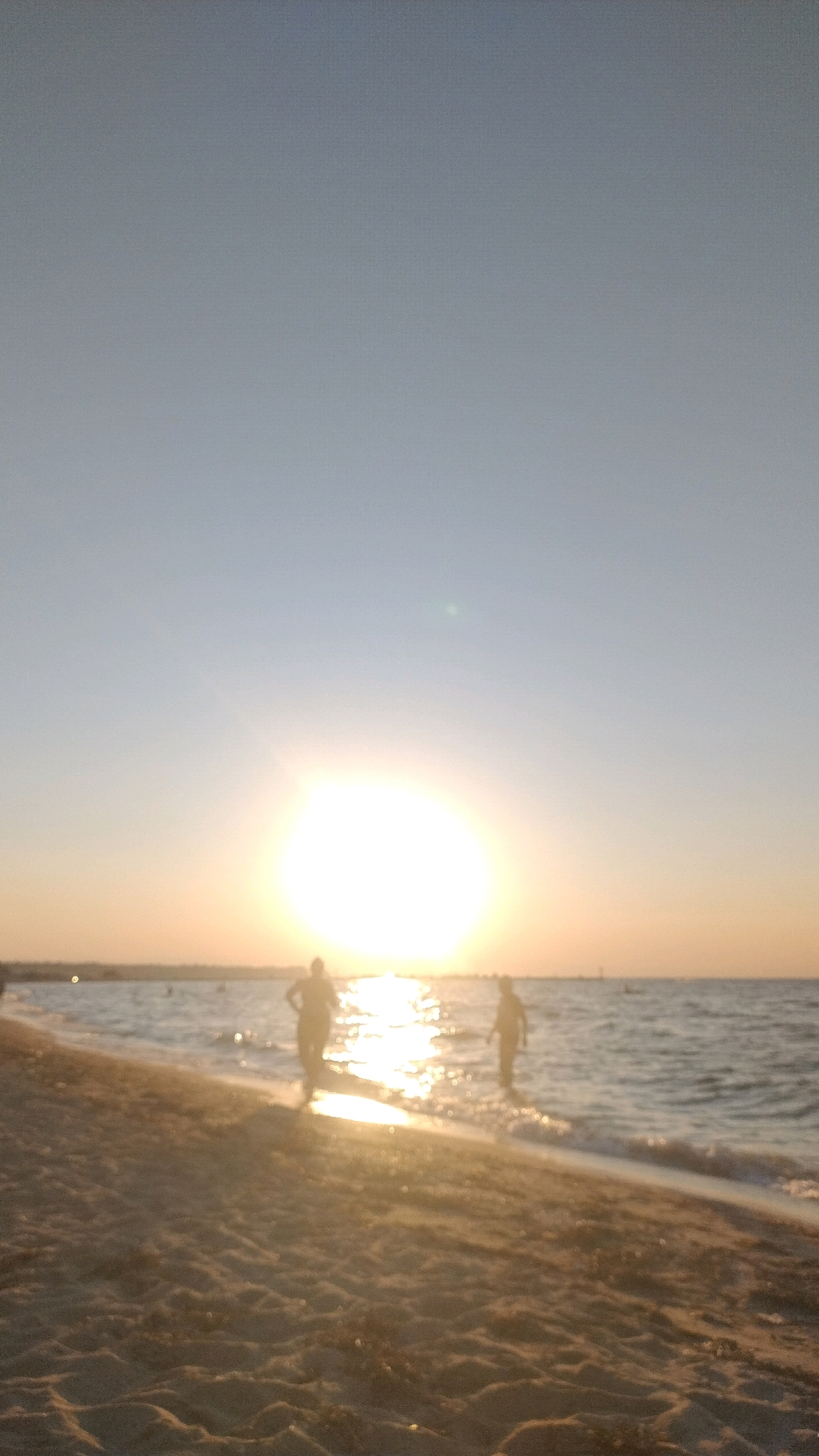 The image depicts a beach scene at Neoi Epivates, Greece, during sunset. The sun is low on the horizon, casting a bright glow and reflecting on the water. Two figures, silhouetted by the sun, are visible near the water's edge. One figure appears to be walking towards the water, while the other is standing in the shallow water. The foreground shows a sandy beach with textured sand. The water is calm with small waves lapping at the shore. The sky is clear and transitions from a light blue at the top to a warm orange hue near the horizon. The overall impression is one of a tranquil evening by the sea.