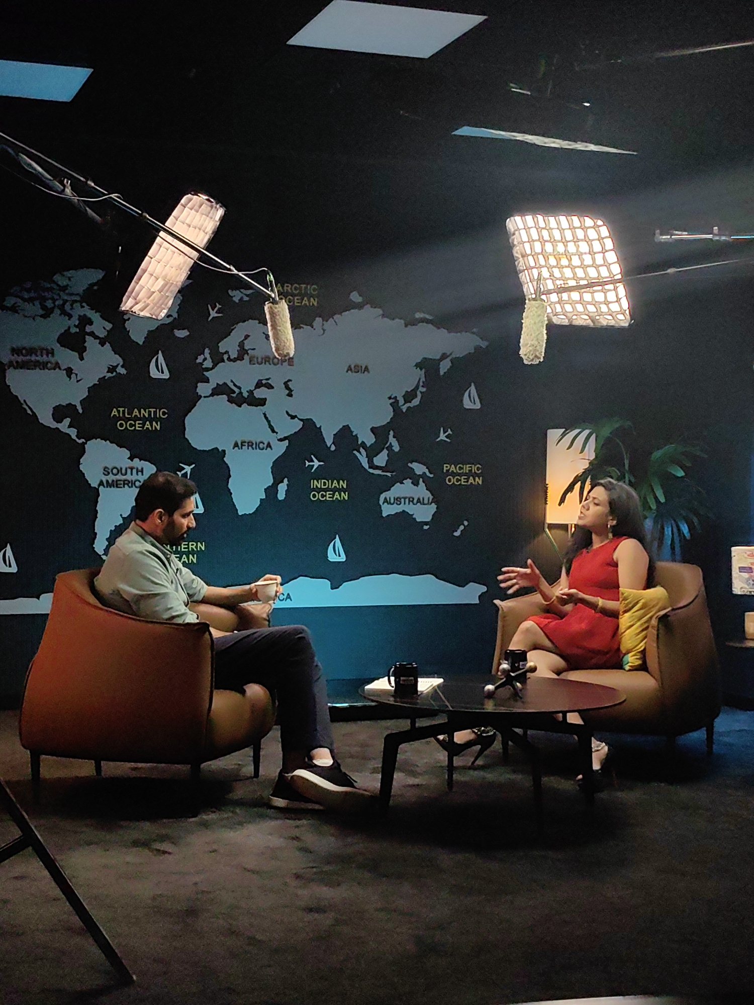The image depicts a studio setting with a large world map as a backdrop. Two individuals are seated in armchairs, engaged in conversation. On the left, a man wearing a light green shirt and dark pants is holding a cup and looking towards the woman. On the right, a woman in a red dress is gesturing with her hands while speaking. A low coffee table is placed between them, with two mugs and a decorative item on it. Two large studio lights are suspended from the ceiling, illuminating the scene. The overall environment appears to be a professionally lit studio space, possibly for a talk show or interview. There is no identifiable information to confirm the location as Mumbai, India, or any specific city.