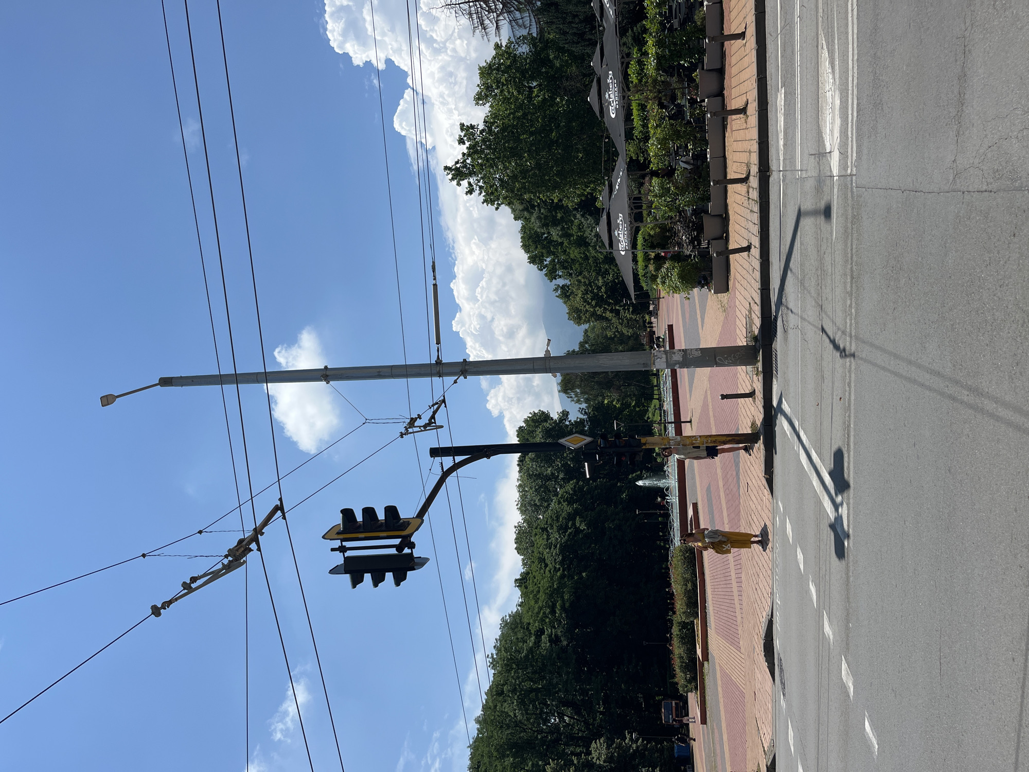 This image depicts an outdoor scene in Sofia, Bulgaria, on a sunny day with scattered clouds. The foreground shows a street with a pedestrian crossing and a sidewalk paved with red and yellow bricks. A traffic light hangs over the intersection, along with overhead power lines, suggesting the presence of public transportation like trams.
On the sidewalk, a person wearing a yellow dress stands near a street lamp, casting a shadow on the road. Further down the sidewalk, a few other individuals are visible, including a person by a fountain and some seated at an outdoor cafe with black umbrellas. Lush green trees and foliage line the edge of the sidewalk, creating a park-like atmosphere. The street itself is paved and shows some signs of wear. The presence of a bus in the background indicates an urban environment. The time of day appears to be daytime, given the bright sunlight and clear shadows. The overall impression is a pleasant, possibly warm, day in a city with green spaces.