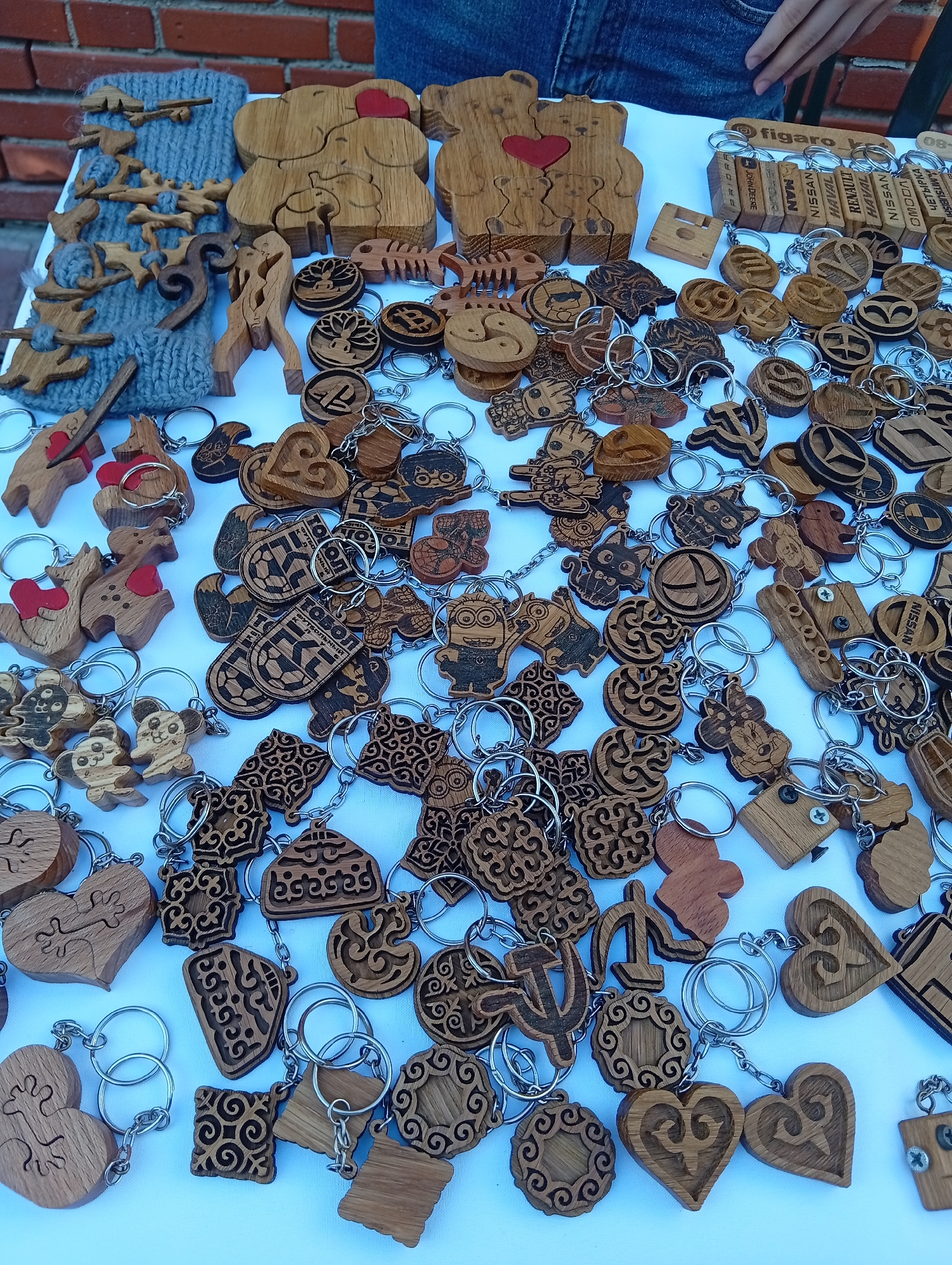 The image depicts a close-up view of a table covered with a white cloth, laden with numerous wooden keychains and wooden decorative items. The keychains are predominantly made of light-colored wood and feature a variety of carved designs, including animals (bears, deer, cats, mice), geometric patterns, symbols (hammer and sickle, dollar sign, Yin Yang, Bitcoin logo), and stylized human figures. Many of these wooden pieces are attached to metal key rings.
In the background, a brick wall is visible, suggesting an outdoor or semi-outdoor setting. Part of a person's legs, clad in blue denim jeans, is also visible at the top of the image. A hand is partially visible reaching from the top right. The lighting suggests it is daytime.
The presence of a person's legs and the arrangement of goods on a table imply a market stall or a craft fair.
Given the prompt's request to include the city of Kostanay, Kazakhstan, this image is presumed to be from that location, though no explicit visual cues within the image directly confirm this. The description of the objects themselves is neutral and does not require specific knowledge of Kostanay.