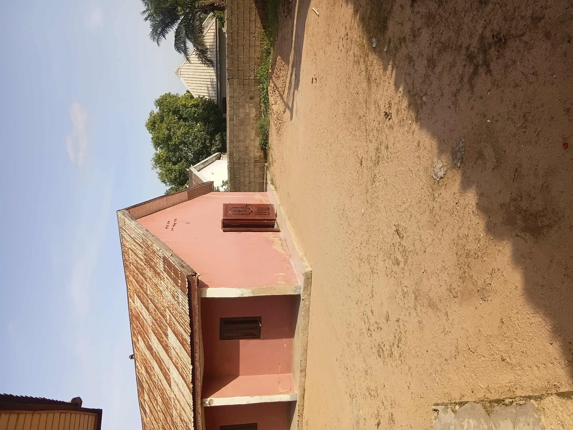 A wide shot captures an outdoor scene in Jalingo, Nigeria, under a clear blue sky with scattered clouds. In the foreground, a sandy, open yard is partially covered by shadows. A pile of sand sits in the middle ground, and a low, grey cinder block wall runs horizontally across the frame. Beyond the wall, several buildings are visible, including a white house with a corrugated metal roof and a palm tree. To the left, a pink building with a rusted corrugated metal roof, dark brown window frames, and an open brown door is prominently featured. The building appears weathered, with peeling paint and a concrete foundation. A partial view of another building's eaves is visible at the top left of the image. The environment suggests a residential or community area in a tropical climate.