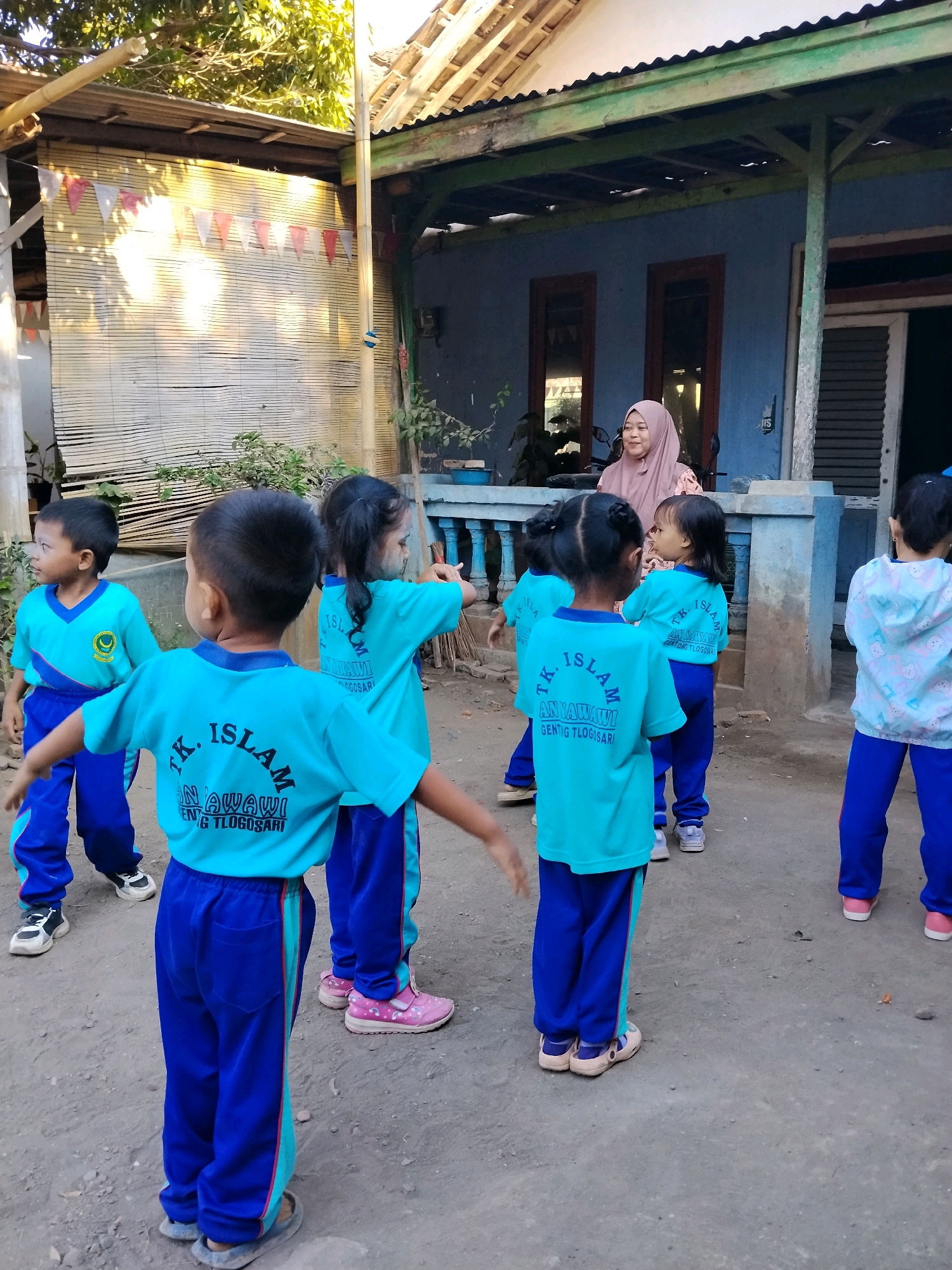 In Panggungan, Indonesia, a group of young children, likely students at TK Islam Annawawi Genting Tlogasari, are gathered outdoors. The children, dressed in matching teal t-shirts with blue pants, appear to be participating in an activity led by an adult woman wearing a pink hijab and patterned shirt, who stands on the porch of a blue-colored building. The environment is rustic, with a bamboo matting wall on the left and a corrugated metal roof overhead. The ground is unpaved, suggesting a simple, functional outdoor space. The children are facing in various directions, with some looking at the adult and others engaged with each other. The overall scene captures a moment of everyday life and education in a local Indonesian community.