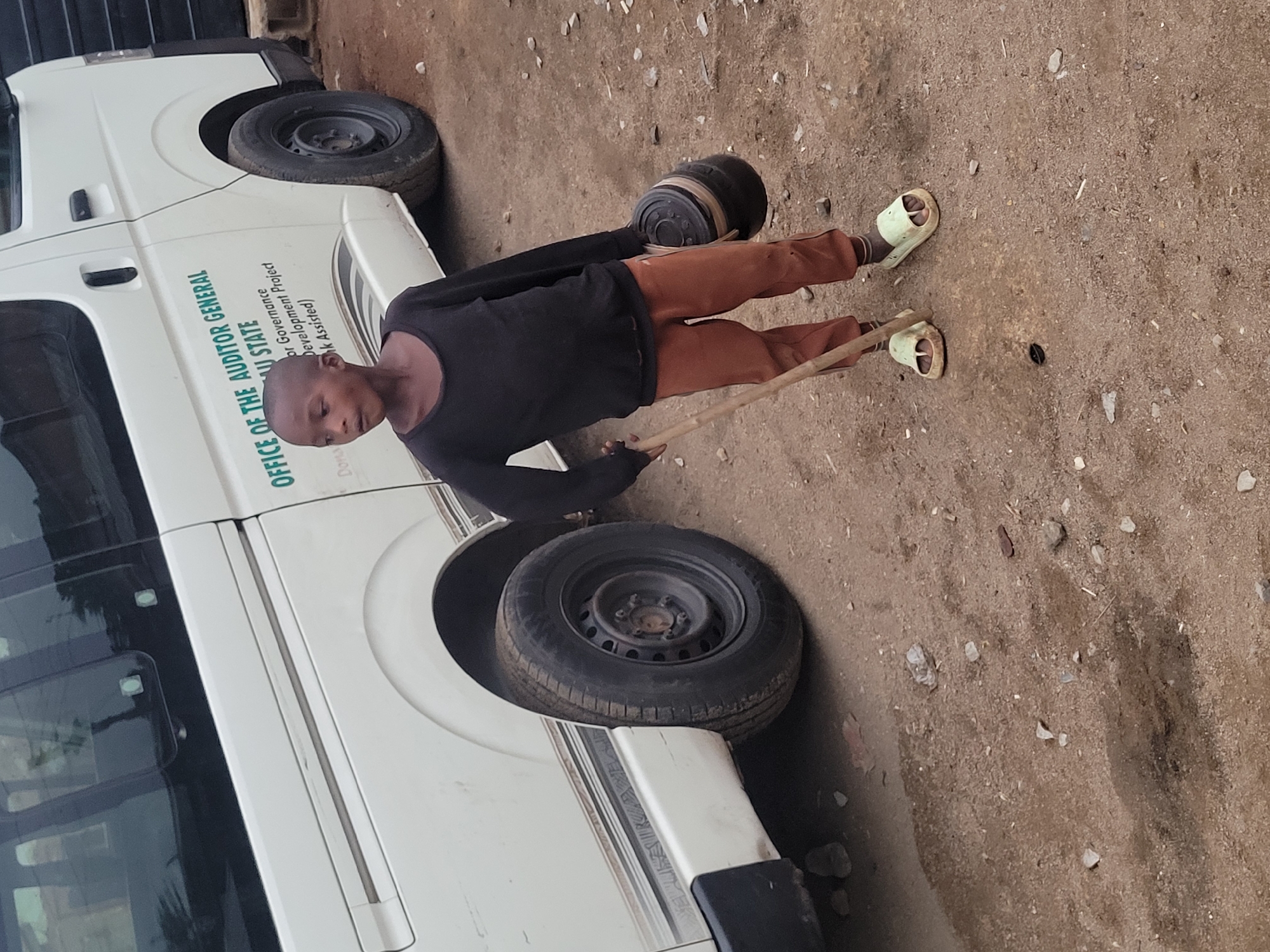 A child wearing brown pants, a black long-sleeved shirt, and light-colored sandals is standing next to a white van. The child is holding a stick in their right hand and appears to be rolling a black tire with a smaller object attached to it. The van has the words "OFFICE OF THE AUDITOR GENERAL" written on its side, followed by the state name and the words "Governance Development Project (World Bank Assisted)." The ground appears to be dirt and gravel. The image was likely taken in Jos, Nigeria, based on the information on the van.