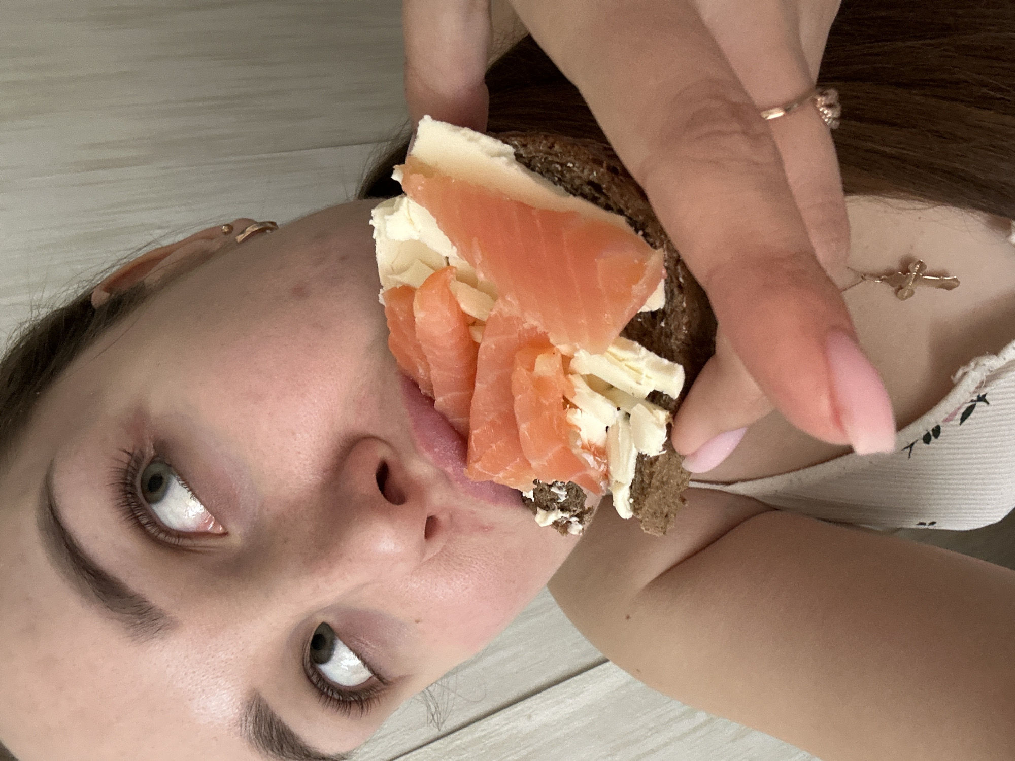 A woman with long brown hair and pale skin is holding a rye bread sandwich with cream cheese and salmon near her mouth. She has a ring on her finger and a necklace. The background appears to be a light-colored wooden surface. The photo has been rotated. There is no information in the image to indicate a location in the city of Chaikovsky, Russia.