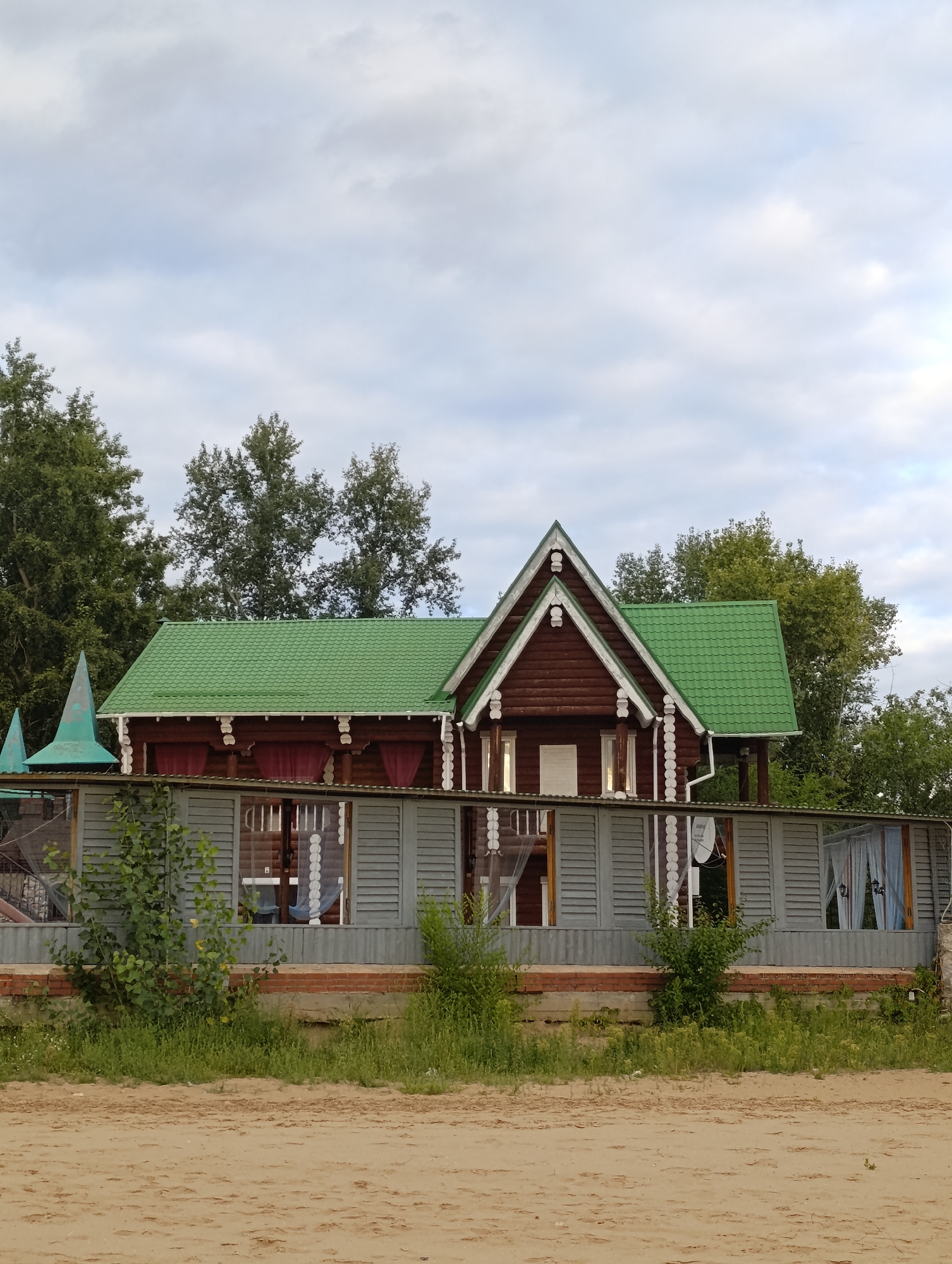 The image shows a wooden house with a green metal roof, located in Tolyatti, Russia. The house has a dark red-brown wooden facade and is decorated with white ornate trim. A gray fence with louvered shutters runs in front of the house. Green trees are visible behind the house and on the sides. The ground in front of the fence is covered with sand and grass. The sky is cloudy. The scene appears to be a residential area, possibly a dacha or a rural property.