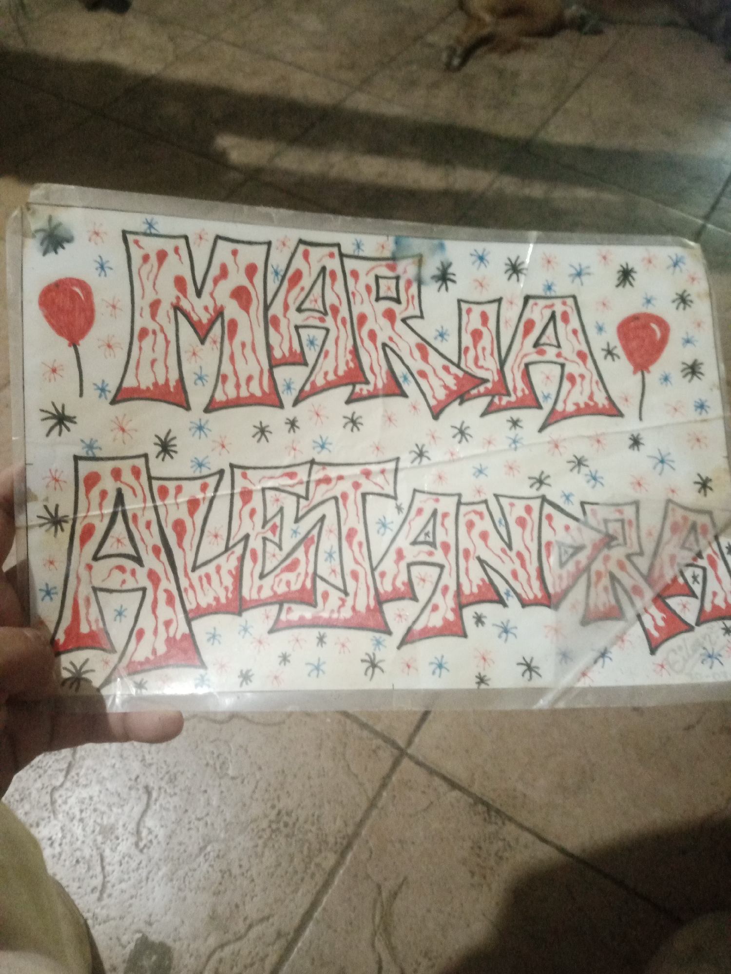The image shows a close-up of a piece of art, likely a banner or sign, with graffiti-style lettering. The lettering is bold and filled with a red and black pattern that resembles dripping blood. The words appear to be "MARIA AVETANA". The background of the art is white and decorated with small, star-like shapes in red, blue, and black, as well as two red balloons. A person's hand is visible on the left side, holding the bottom edge of the artwork. The artwork is placed on a tiled surface, possibly a floor or wall, in what appears to be an indoor setting. The overall lighting is dim. There is no specific information in the image to indicate its location in the city of Santa Ana, Venezuela.