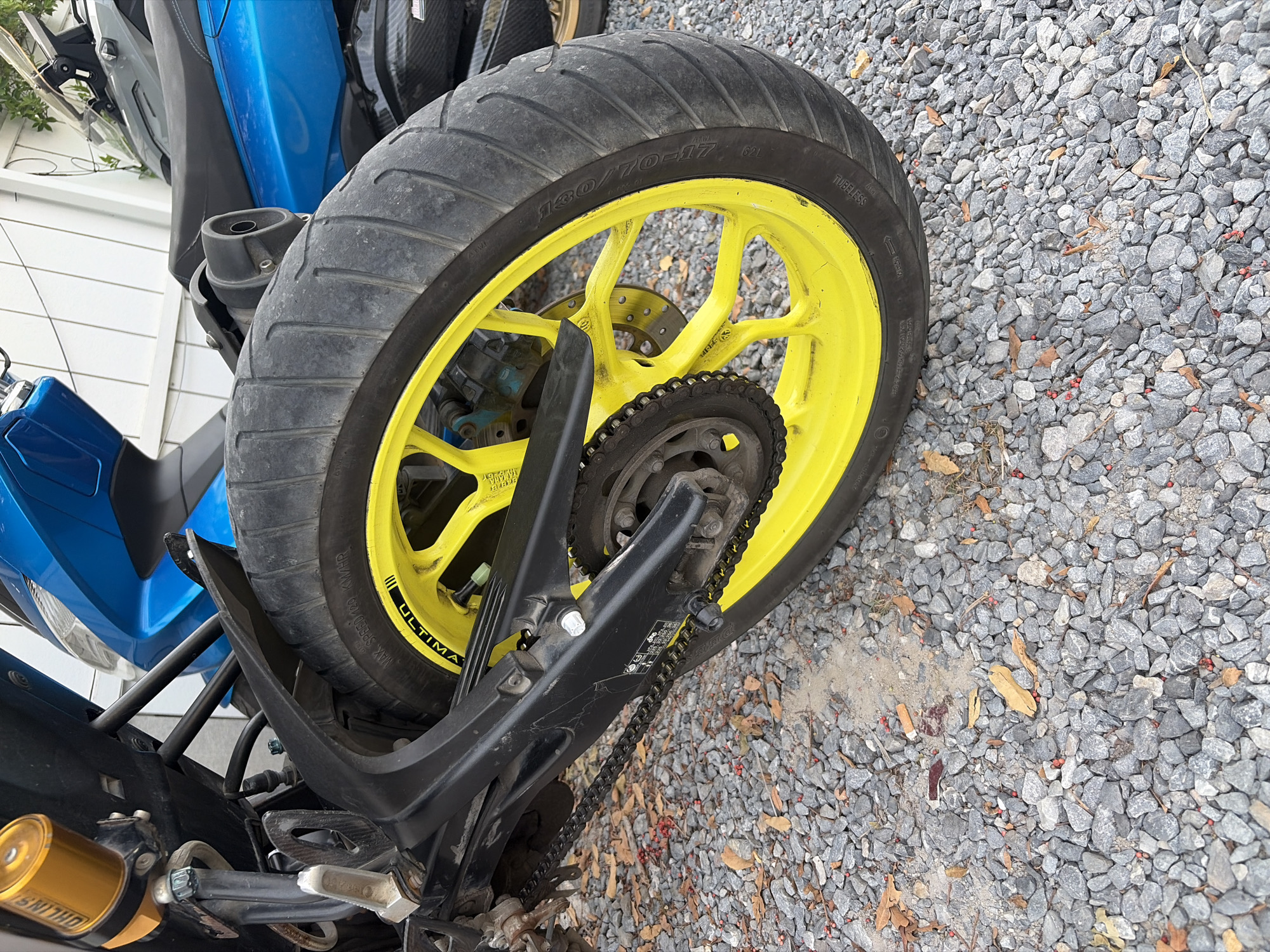 This media file shows the rear wheel and chain of a blue and black motorcycle. The motorcycle has a bright yellow rim and a black tire with "130/70-17" and "TUBELESS" written on it. A black chain connects the rear sprocket to the engine, which is not fully visible. The motorcycle is parked on a gravel surface with some scattered leaves and small red berries. In the background, a portion of a white-paneled building and another motorcycle, also predominantly blue, can be seen. The lighting suggests it is daytime. The scene appears to be an outdoor setting, possibly a residential area or a parking lot. No people are visible in the image. The overall impression is that of a parked motorcycle, possibly in Thailand given the location context, in a simple outdoor environment.