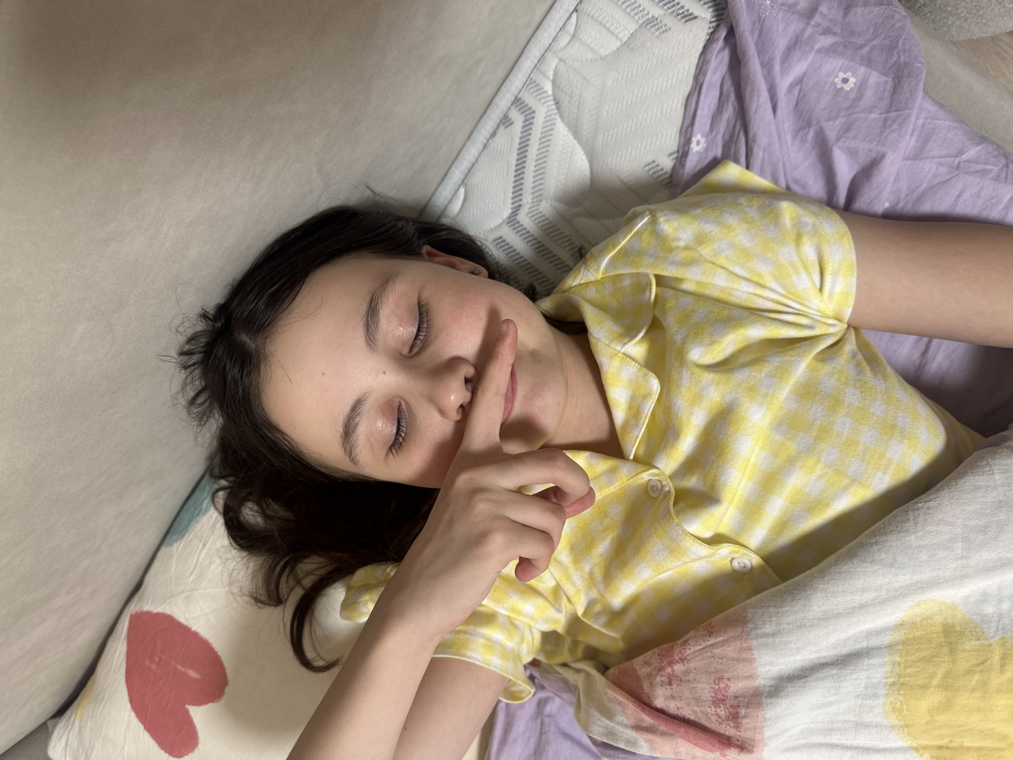 A young girl lies in bed, her eyes closed, with her index finger raised to her lips in a "shh" gesture. She is wearing a yellow and white checkered pajama top. She is lying on a pillow with a red heart on it, and a white mattress. The bed has purple floral sheets. No information indicates a specific location in Votkinsk, Russia.