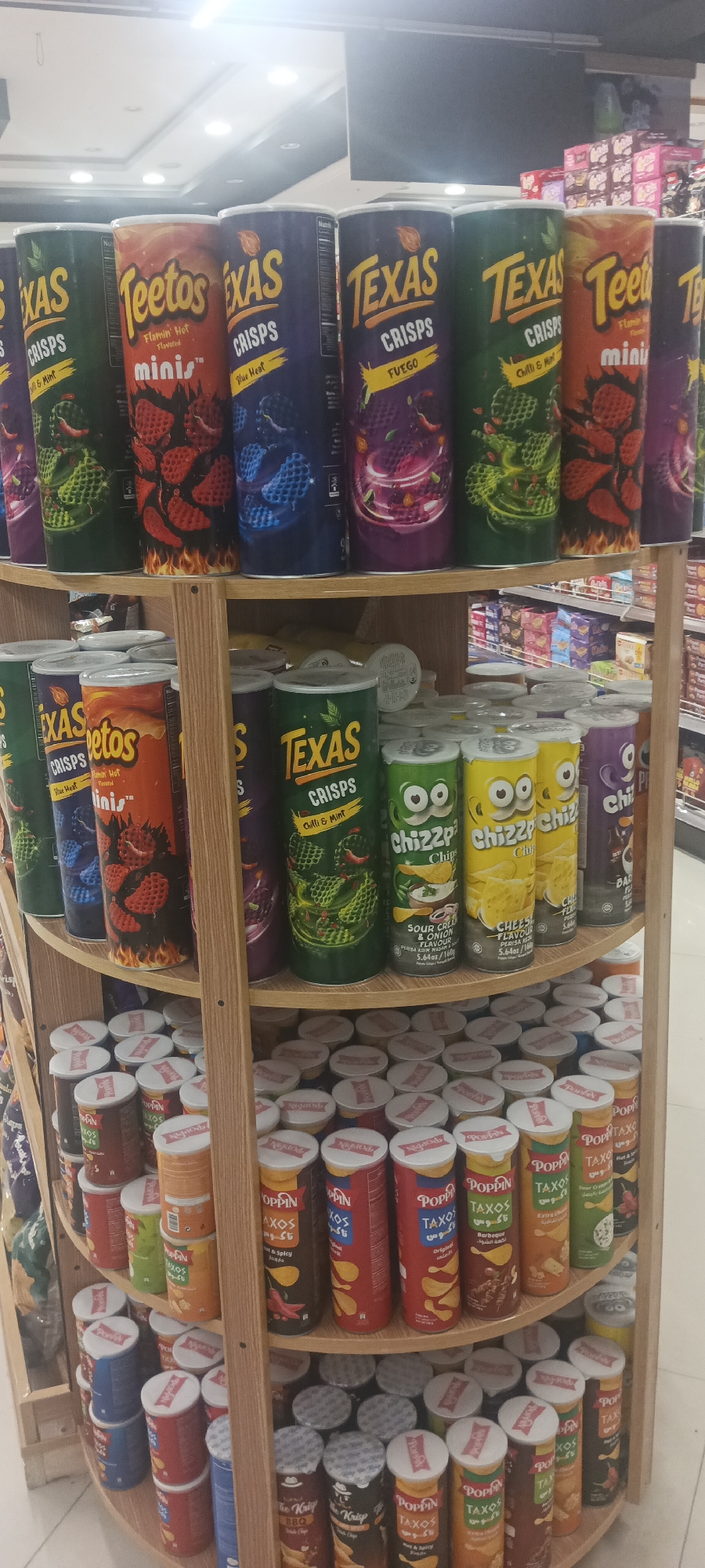 The image shows a retail display of potato chips in a store. The display is on a multi-tiered wooden shelf. The top shelf features several tall cylindrical containers of chips, including brands like "Teetos" and "Texas Crisps" in various flavors such as "mini's," "Flamin' Hot," "Blue Heat," and "Fuego." Some containers have graphics of chili peppers and other spicy ingredients. Below this, on the second shelf, are more cylindrical containers of chips, including "Texas Crisps" and "Chizzpp Chips" with flavors like "Chilli & Mint," "Sour Cream & Onion," and "Cheese." The bottom two shelves are filled with numerous smaller cylindrical containers of chips, mostly with white lids. These smaller containers include brands like "Poppin Taxos" and others with Arabic script. The background shows store shelving with more packaged goods, suggesting a supermarket or convenience store environment. The overall impression is a retail setting offering a variety of snack food products, specifically potato chips, in a range of sizes and flavors. The location is stated to be in Peshawar City Tehsil, Pakistan.