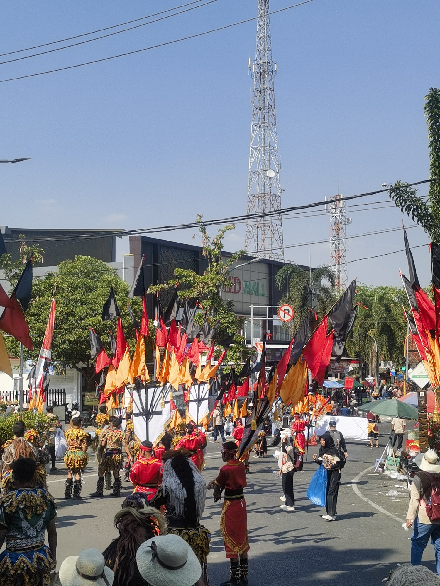 The image depicts a vibrant street parade in Blora, Indonesia, on a sunny day. The foreground is dominated by people in traditional, colorful costumes, some with elaborate headdresses and makeup. Many are holding flags in shades of red, black, and orange, adding to the festive atmosphere. In the background, a tall telecommunications tower stands against a clear blue sky, a stark contrast to the cultural display. Buildings, including a structure with "RED MALL" visible, line the street, along with trees and power lines. A traffic sign with a red circle and a diagonal line indicates no parking. The overall scene suggests a lively community event or cultural festival taking place in an urban setting.