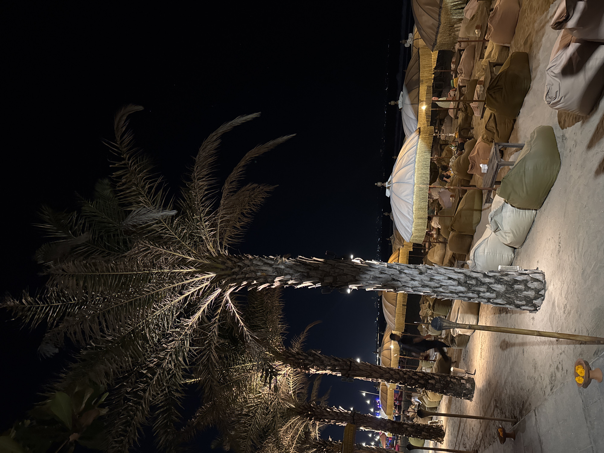 The image depicts a beachside lounge area at night in Kuta, Indonesia, characterized by palm trees, beanbag chairs, and umbrellas. Several palm trees, illuminated by spotlights from below, dominate the foreground and midground, their fronds casting shadows. The ground is sandy, with a paved walkway visible on the left.
Scattered across the sand are numerous large, beige and olive-green beanbag chairs, some occupied by people. Small wooden tables are placed between the beanbags. Overhead, large, beige umbrellas with gold fringe provide shade, some illuminated from within. A few individuals are visible, including a person walking between palm trees in the midground and others seated in the lounge area.
The background shows the dark expanse of the ocean and a distant, faintly lit cityscape. The overall atmosphere is dimly lit, with artificial lighting highlighting the seating area and palm trees against the dark night sky.