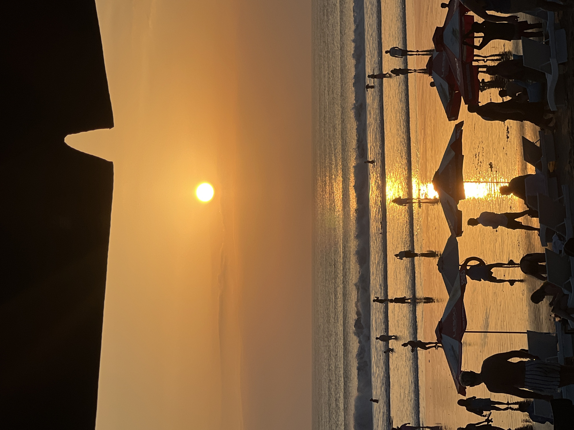 A sunset scene on Kuta Beach, Indonesia, is depicted, dominated by a warm, golden-orange sky. The sun, a bright disc, is visible in the upper left quadrant, casting a shimmering reflection on the ocean. Gentle waves roll towards the shore, where numerous silhouetted figures are scattered. Some individuals are standing in the shallow water, while others are closer to the beach. In the foreground, beach umbrellas cast shadows, and lounge chairs are visible. The overall atmosphere is serene and highlights a typical evening at this popular tourist destination.