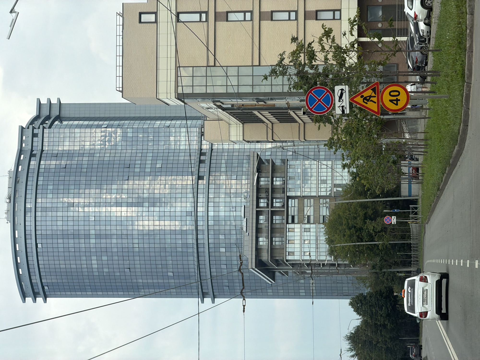 The image captures a street view in Kazan, Russia, dominated by modern architecture. A tall, cylindrical building with a glass facade stands prominently in the background, reflecting the sky and surrounding structures. To its right, a multi-story brick building with large glass windows is visible. In the foreground, a white taxi with "Яндекс Go" branding is driving down a road. Several traffic signs are present on the right side of the road, including a "No Parking" sign, a "Tow Away Zone" sign, a "Road Works Ahead" sign, and a speed limit sign indicating 40 km/h. A row of young trees lines the sidewalk. The sky is overcast, and the overall scene suggests an urban environment.