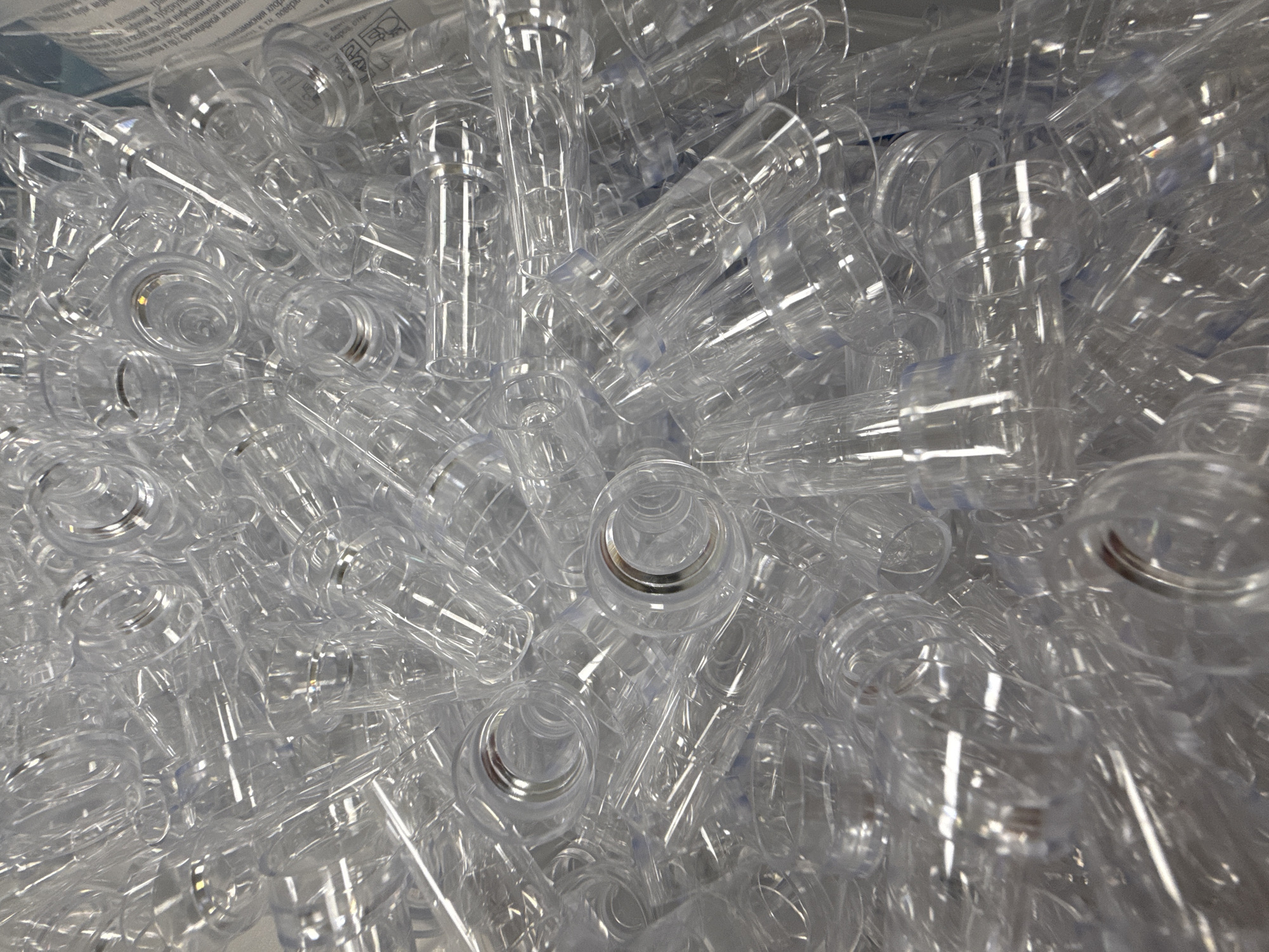 A densely packed collection of clear, hollow, plastic objects with cylindrical and conical shapes are filling the frame, suggesting they might be preforms for plastic bottles or similar containers. The lighting is bright and the focus is sharp, revealing the translucence and smooth surfaces of the objects. There is a small section of printed material with text in Russian visible in the upper left corner of the frame. The text reads "и особо опасных инфекций пошения вирусов полиомиели грипа и пр. фунгицидной активн Бензиламмония хлор ти повер боров, отра". There are no indications of the city of Moscow or people present in the frame.