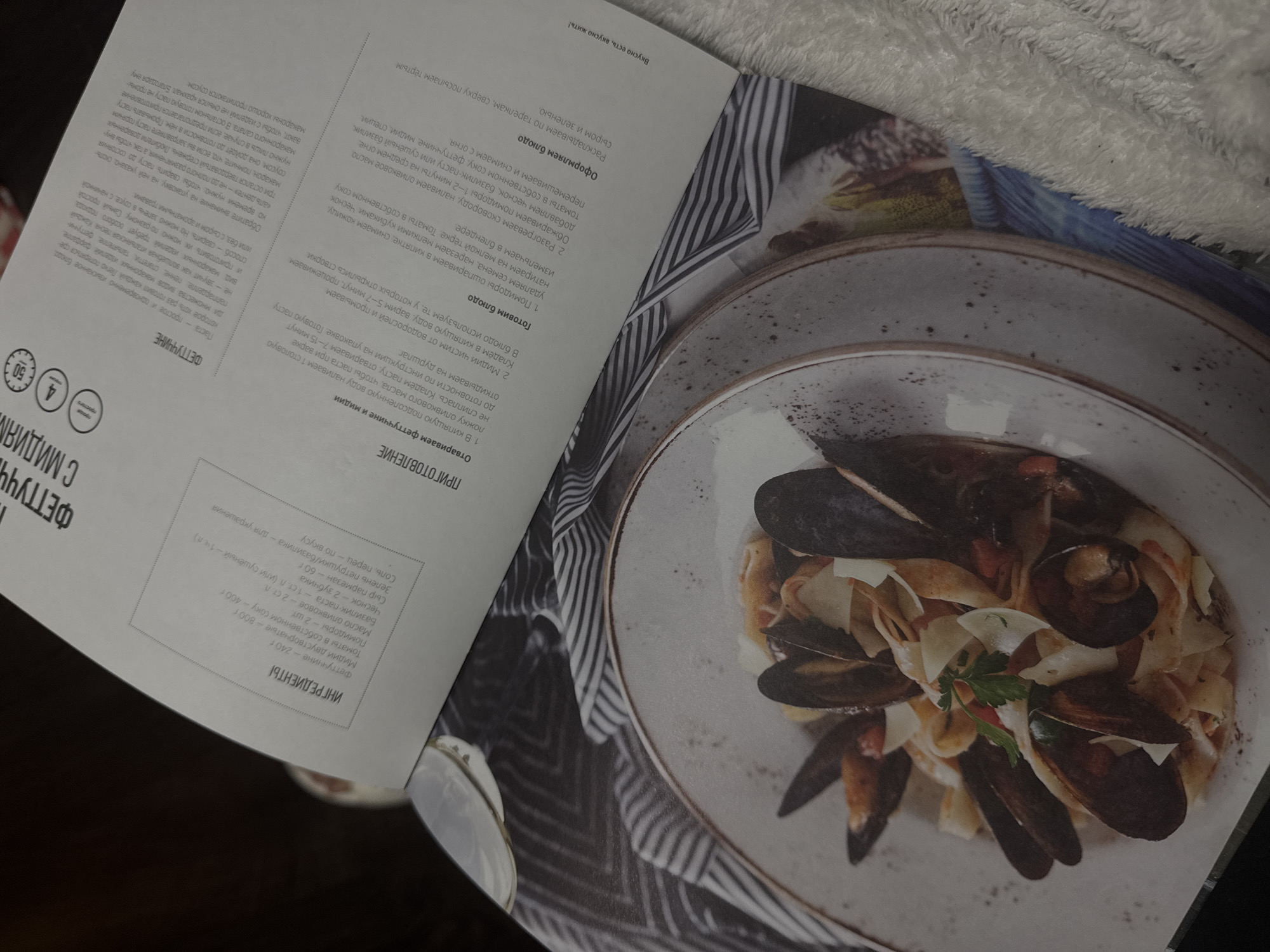 The image shows an open cookbook with a recipe and a photo of fettuccine with mussels. The page on the left lists the ingredients: fettuccine, tomatoes, mussels, canned tomatoes, olive oil, basil paste, garlic, Parmesan cheese, salt, pepper, and parsley/basil. The page on the right provides the cooking instructions, which include boiling the fettuccine and mussels, preparing the sauce with the tomatoes and seasonings, and assembling the dish. A plate of fettuccine with mussels, garnished with herbs and cheese, is shown in the photo. The background includes a striped fabric. The location in Izhevsk, Russia is not discernible.