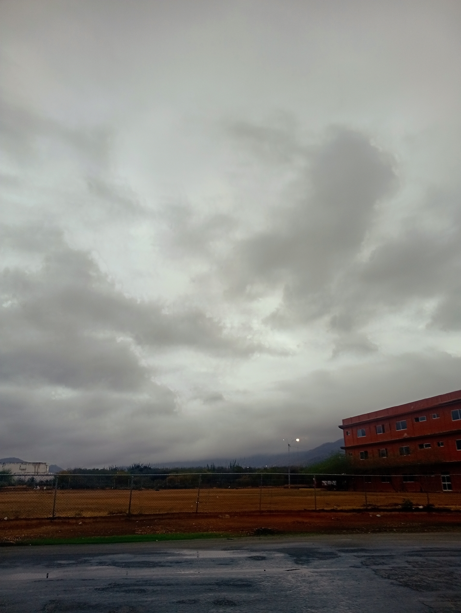 The image depicts an overcast sky over a scene in Juan Griego, Venezuela. In the foreground, a wet asphalt surface reflects the dim light. A chain-link fence runs horizontally across the mid-ground, separating a patch of dry, brown earth from what appears to be a field or undeveloped land behind it. In the distance, a multi-story, reddish-orange building with numerous windows is visible on the right side of the frame. To the left of the building and further in the background, a low-lying structure with white siding is present, with hills and mountains forming a backdrop. The overall atmosphere is muted and somber, suggesting either pre-storm conditions or the aftermath of rain. No people or animals are visible in the frame.