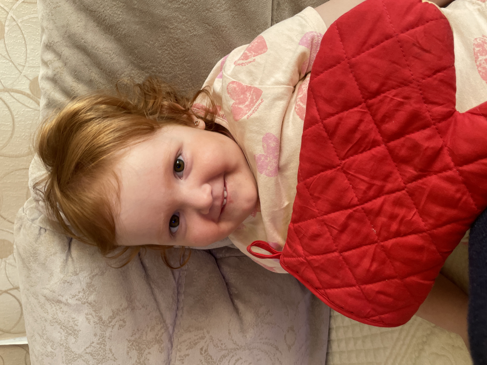 A young child with reddish-brown hair is lying on a beige pillow. The child is wearing a light-colored shirt with pink heart patterns. A red oven mitt is positioned near the child's body. The location is not discernible from the image and is therefore not identifiable as Кипчак, Russia.