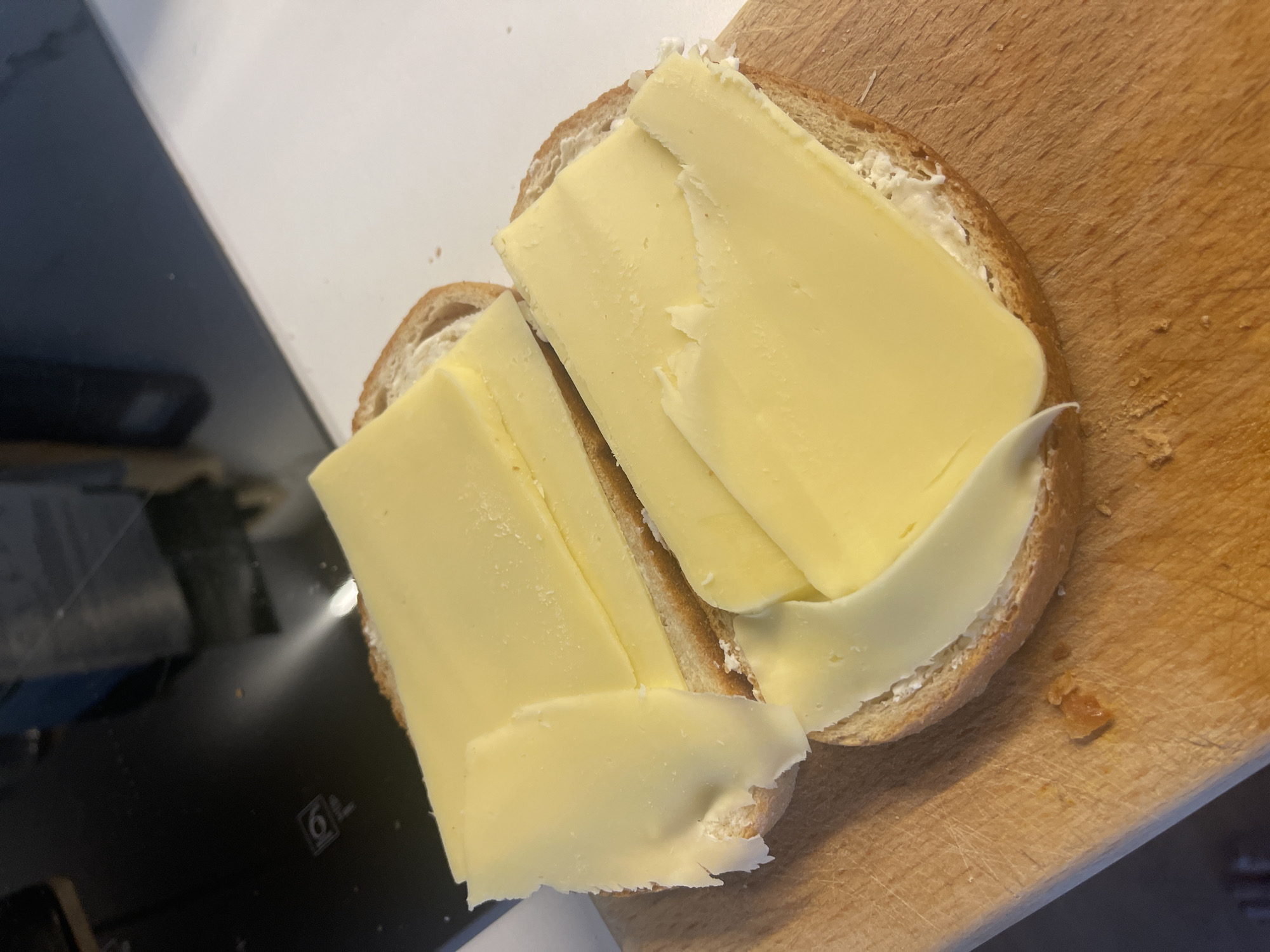 The image shows two slices of bread topped with sliced cheese on a wooden cutting board. A white countertop and a black appliance are visible in the background. The scene is indoors.