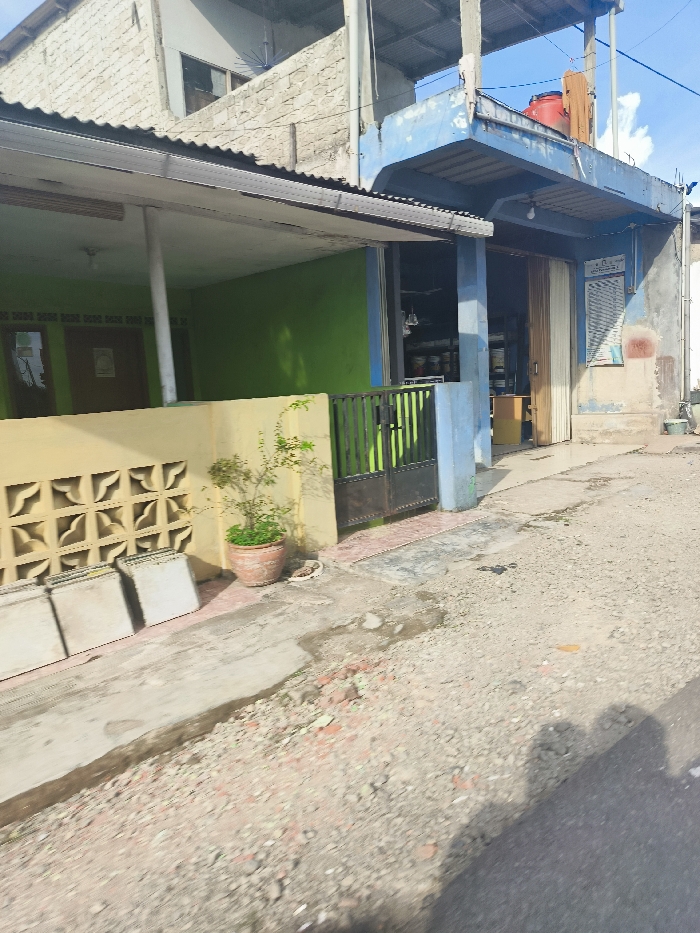 The image shows a street scene in Sukatani, Indonesia. The scene is comprised of two buildings, one in front of the other. The front building is a single story structure with a light blue facade, a green door, a black metal gate and a yellow wall with a concrete grid patterned section. The back building is a two story structure with a gray and beige facade. The street is paved with gravel and has a shadow stretching across it. A small plant in a terra cotta pot is located in front of the yellow wall. There are no people present.