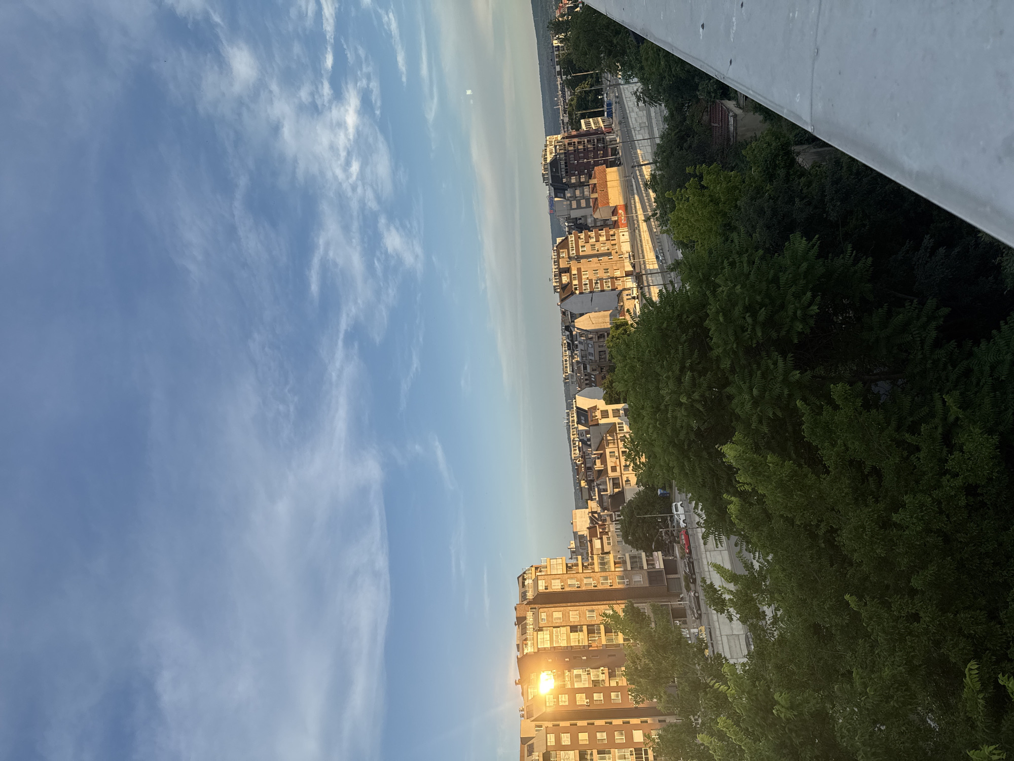 The image captures a cityscape during the golden hour, likely late afternoon or early evening, given the warm lighting on the buildings and the sky. The scene is dominated by several mid-rise apartment buildings with balconies, their facades illuminated by the setting sun on the left side of the frame. In the foreground and mid-ground, lush green trees provide a vibrant contrast to the urban architecture. A road or elevated pathway can be seen behind the trees, with a few cars driving on it. The sky is a clear blue, interspersed with wispy white clouds, suggesting fair weather. The overall impression is one of a typical urban environment bathed in the soft glow of sunset.