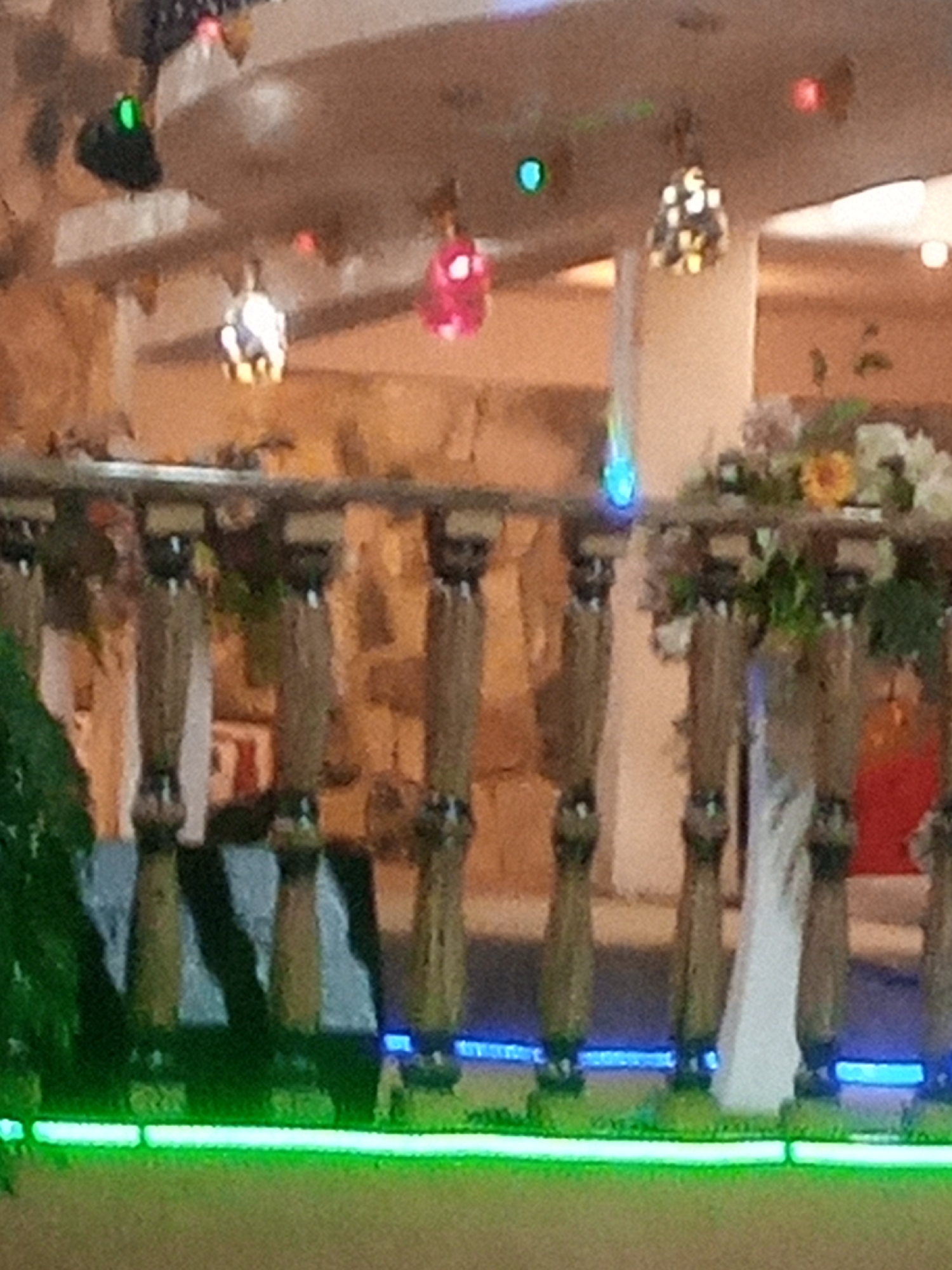 The image depicts a dimly lit interior with a textured, stone-like wall in the background. In the foreground, there is a wooden railing with decorative pillars. Green and blue LED lights are visible along the base of the railing. Several decorative lamps and what appear to be artificial flowers are arranged on a shelf above the railing. The overall scene suggests an indoor decorative setting, possibly a restaurant or a similar establishment, located on Rukuba Road, Nigeria. There are no people visible in the image.