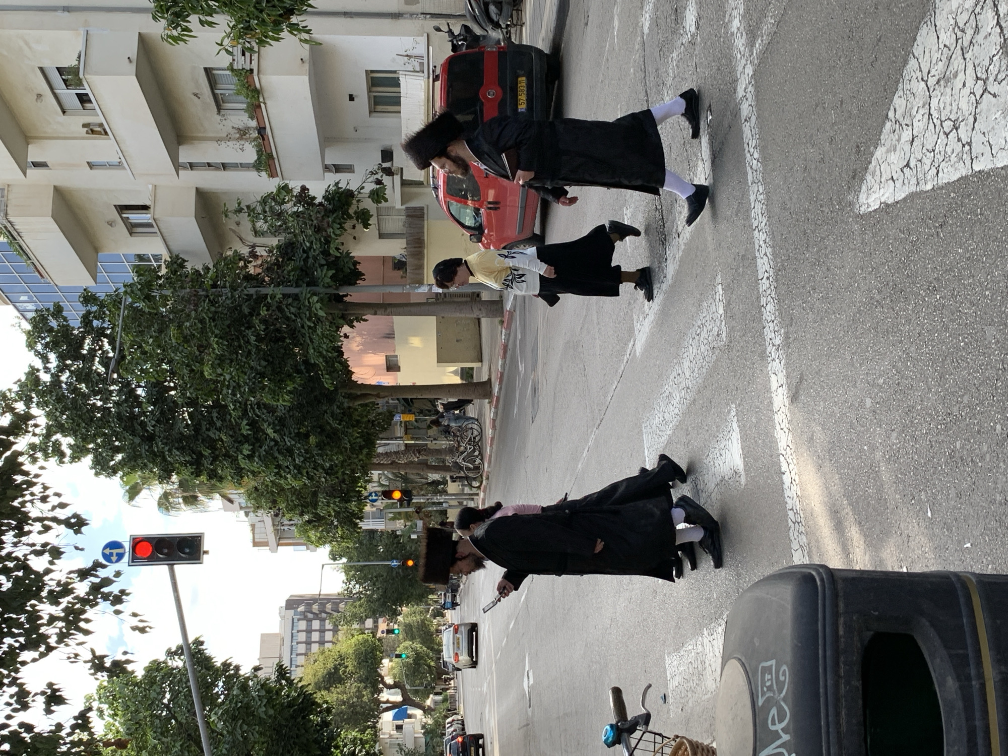 The image captures a street scene in Tel Aviv, Israel, with three Orthodox Jewish men in traditional attire—black coats and hats, and white socks—crossing a pedestrian crosswalk. A traffic light overhead is red for vehicular traffic. The street is lined with buildings, including residential and commercial structures. There's greenery in the form of trees and shrubs along the street. A black trash bin and a bicycle are in the foreground. A red car is parked on the side of the road.