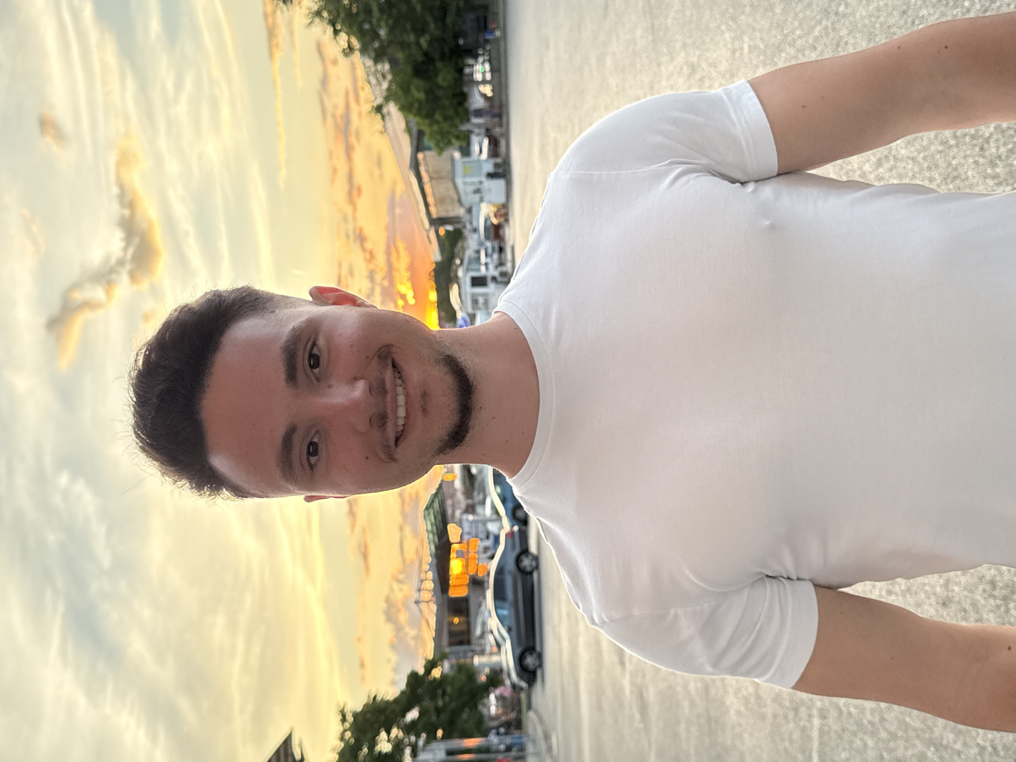 A young man with short, dark hair and a beard is standing in an outdoor parking lot during sunset. He is wearing a white t-shirt and smiling at the camera. The background shows a street with cars, buildings, and trees, all illuminated by the warm glow of the setting sun. The sky is filled with colorful clouds, ranging from yellow to orange and pink. The man is positioned in the foreground, with the parking lot and the street scene behind him. The image appears to have been taken in the city of Sofia, Bulgaria, based on visual cues in the background.
