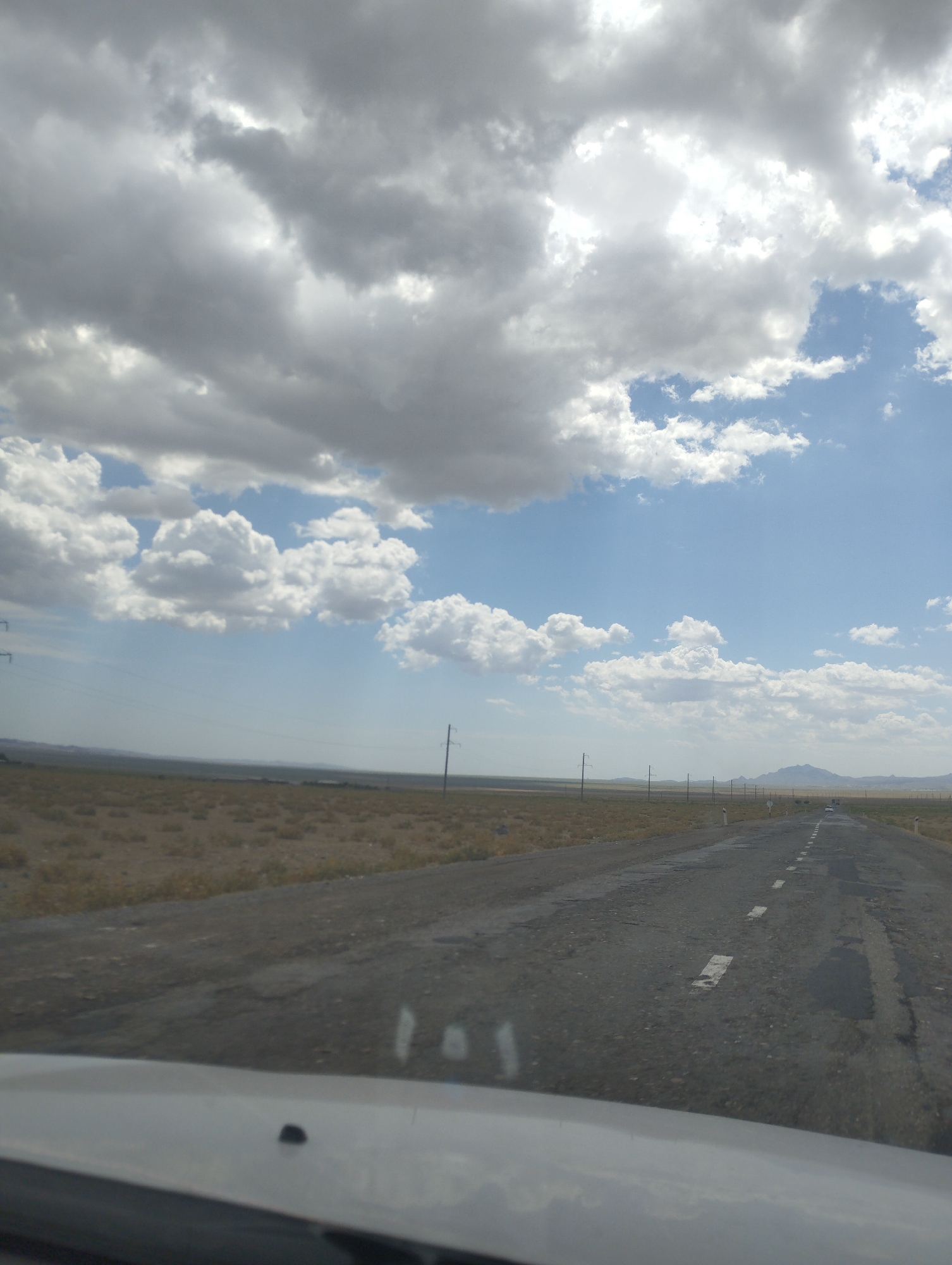The image shows a view from a car driving on a long, paved road that stretches into the distance under a dramatic sky filled with clouds. The road is cracked and weathered, with faded white lines marking the lanes. To the left and right of the road, the landscape is flat and arid, covered in dry, sparse vegetation. In the far distance, the land rises into rolling hills and mountains, suggesting a rural or semi-desert environment. Several utility poles are visible along the roadside, indicating infrastructure. A few vehicles can be seen in the distance, also traveling on the road. The sky is a mix of bright blue patches and large, dense grey and white clouds, with sunlight breaking through in places, creating a dynamic play of light and shadow. The location is identified as being in the Forish QFY, Uzbekistan.