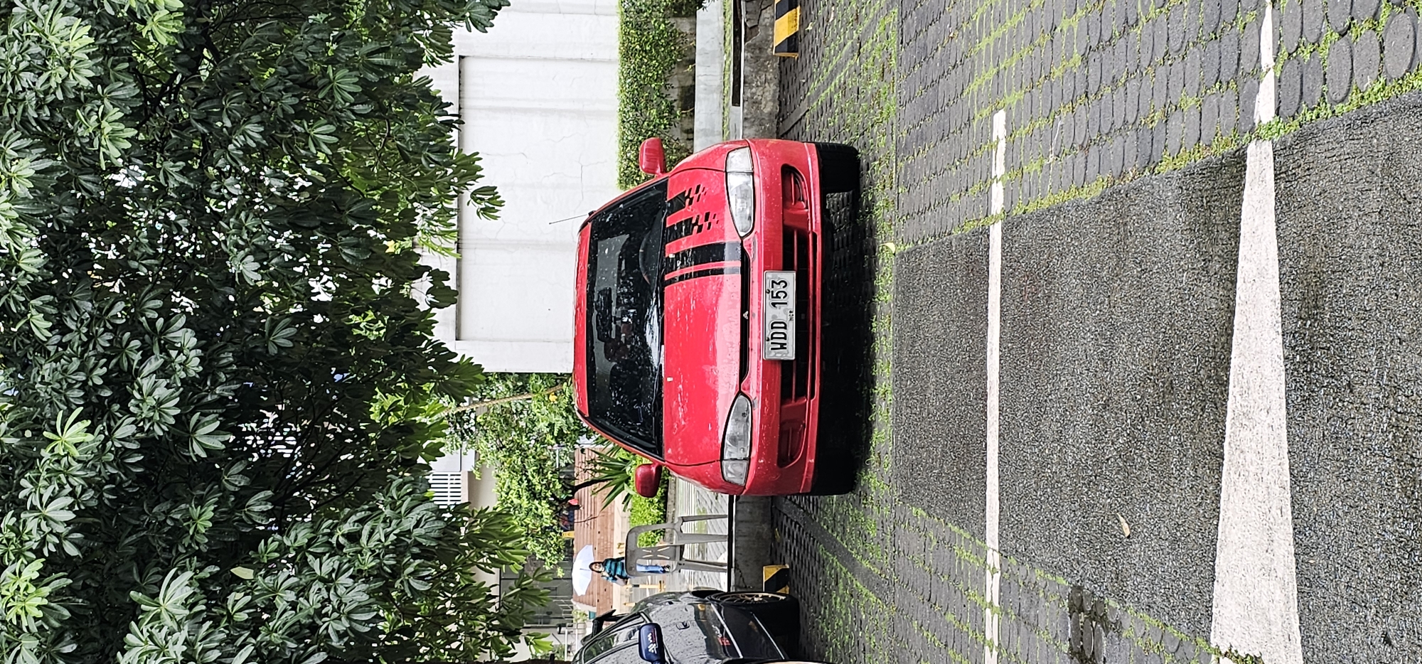 The image captures a red car parked on a patterned paved lot in Quezon City, Philippines, under the canopy of a large tree. The car, a Mitsubishi Lancer, is the central focus, with racing stripes on its hood and a license plate reading "WDD 153 NCR." In the background, a woman holding a white umbrella walks away from the car toward a building with stairs. The scene appears to be a parking area in front of a building, likely an office or residential complex, as suggested by the trees, paved lot, and surrounding architecture. The ground is wet, indicating recent rainfall.