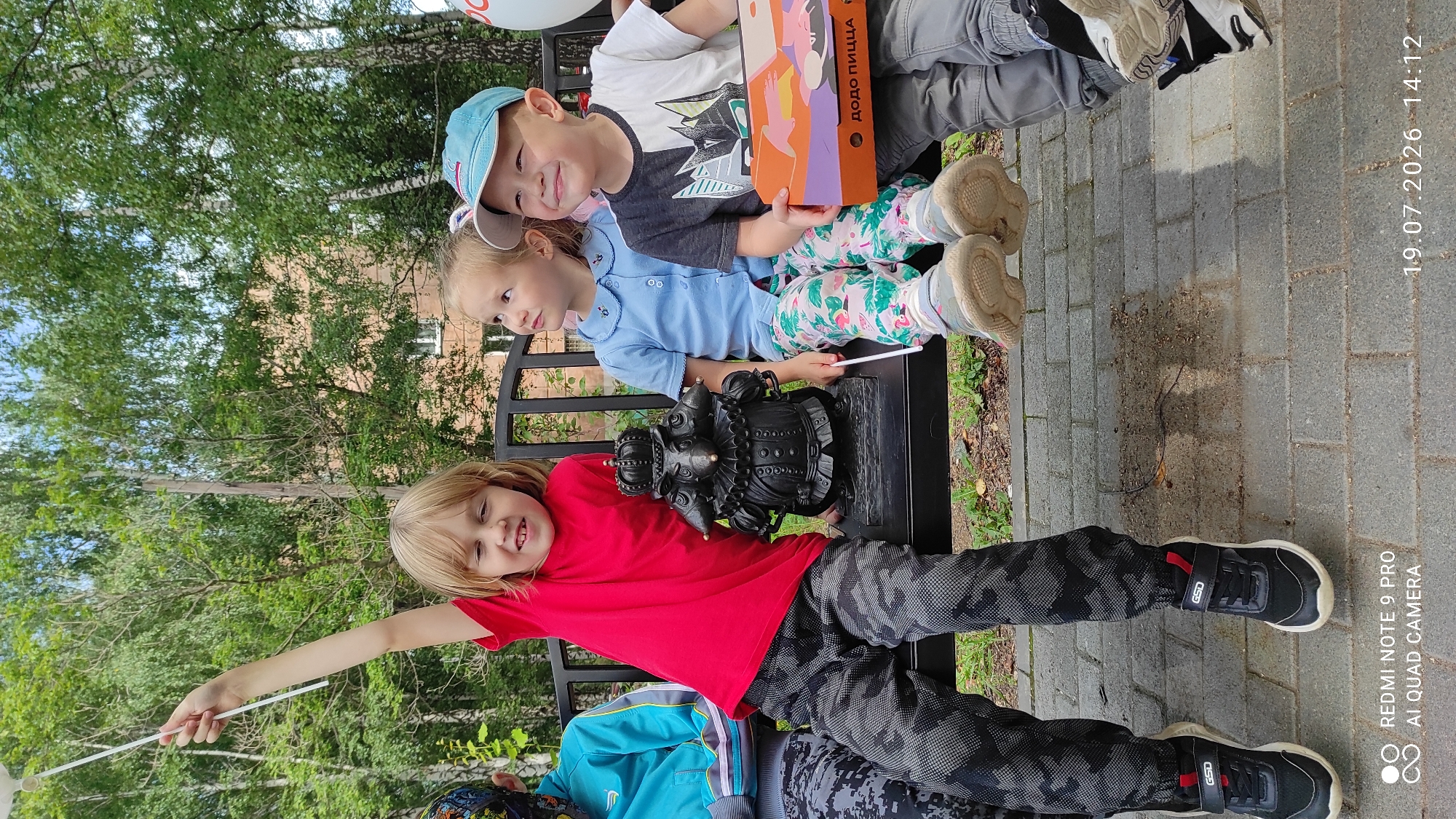 The image depicts three young children seated on a park bench outdoors, with a small, dark, statue of a creature wearing a crown and ruff between two of them. The child on the left, a girl with blonde hair, is wearing a red t-shirt and camouflage pants, and is holding a white wand-like object aloft. The child in the middle, a girl with light brown hair, is wearing a blue collared shirt and patterned pants. The child on the right, a boy wearing a blue cap, a black and white t-shirt, and gray pants, is holding an open orange pizza box with Russian text that appears to say "ДОДО ПИЦЦА". They are all looking upwards and to the right, with expressions of curiosity or amusement. The bench is set on a paved area with a brick-like pattern. In the background, trees with green foliage are visible, and a building with windows can be seen through the trees. The overall scene suggests a casual outdoor gathering, possibly a birthday party given the pizza box. The timestamp "19.07.2026 14:12" is overlaid on the bottom right of the image. The text "REDMI NOTE 9 PRO AI QUAD CAMERA" is overlaid on the bottom left. This is likely a residential area or park in Chaikovsky, Russia, given the Cyrillic text on the pizza box and the typical park scenery.