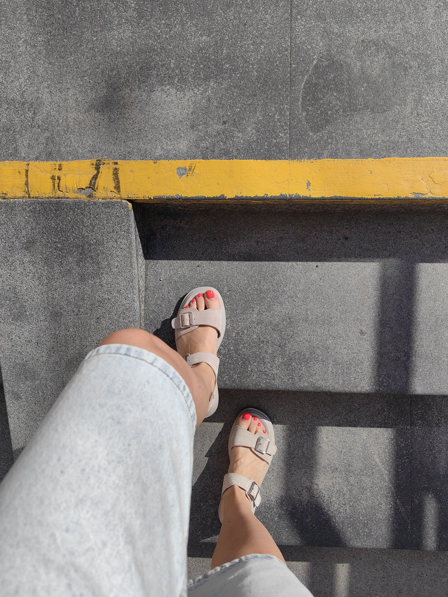A first-person perspective image shows a person standing on grey stone steps with a bright yellow line marking the edge of the top step. The person is wearing a light blue denim skirt and beige sandals with silver buckles. Their toenails are painted a vibrant coral red. Sunlight casts shadows on the steps. The location is an outdoor urban environment.
