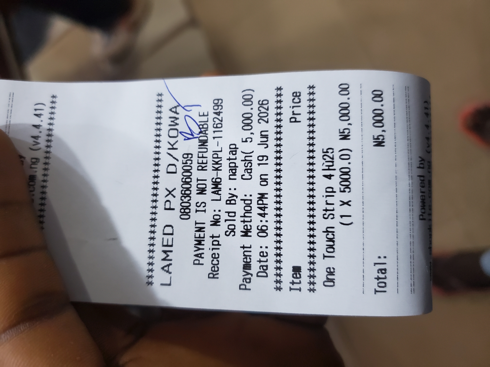 The image shows a close-up of a receipt held by a person's hand. The receipt is for a purchase made at "LAMED PX D/KOWA" with a phone number "08036060059". The transaction details include "PAYMENT IS NOT REFUNDABLE", "Receipt No: LAM6-KKPL-1162499", "Sold By: naptap", "Payment Method: Cash( 5,000.00)", and "Date: 06:44PM on 19 Jun 2026". The item purchased is "One Touch Strip" for N5,000.00, with a total of N5,000.00. The receipt also mentions "Powered by" and a website address. The background suggests an indoor setting with blurred brown tones. The currency symbol "N" indicates Nigerian Naira. While the image does not explicitly state the location in Jos, Nigeria, the presence of Nigerian currency and the typical format of receipts suggest a transaction within Nigeria.