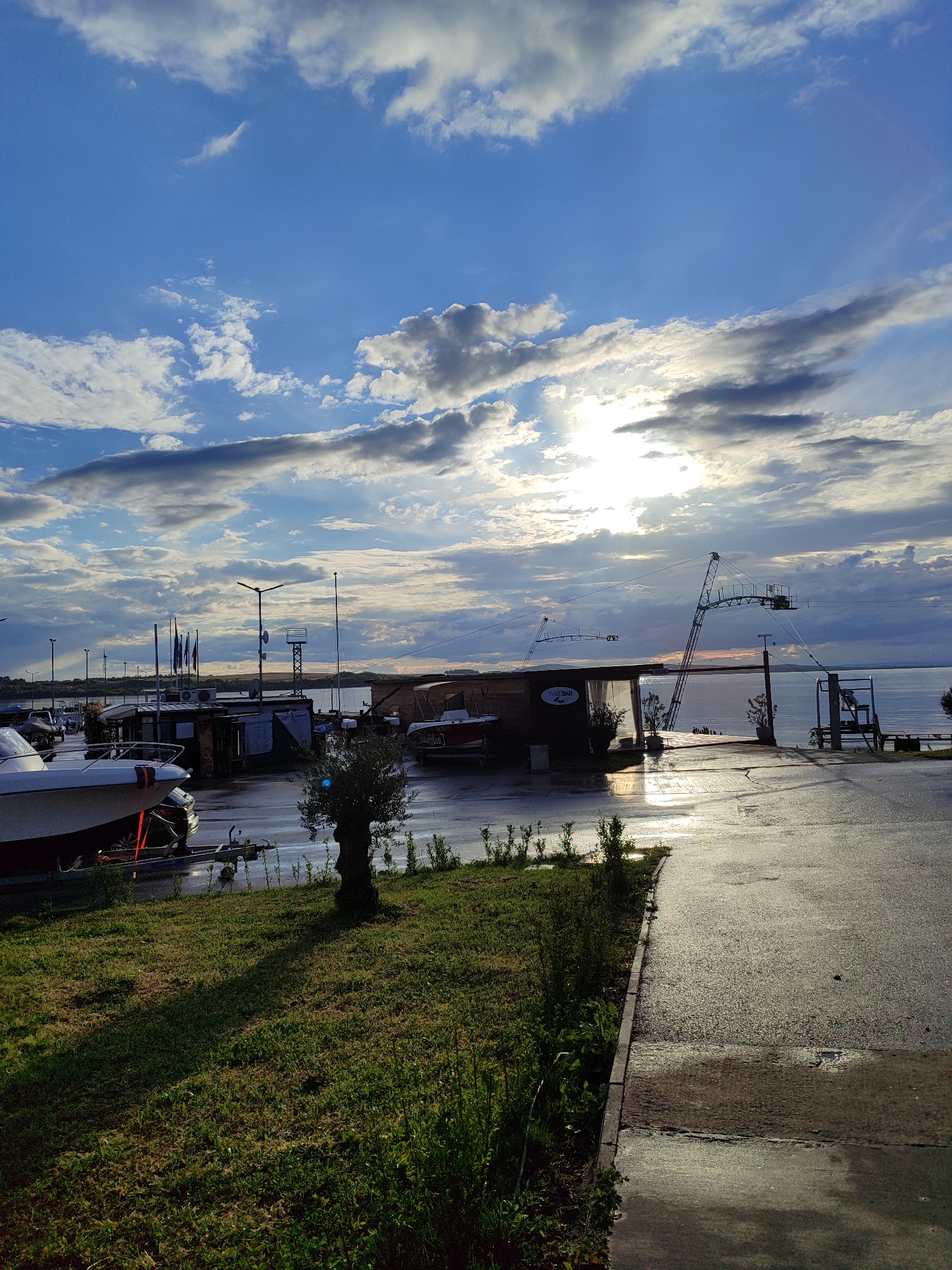 The image depicts a marina scene on a wet, paved area with a grassy foreground. The sky is predominantly blue with scattered white and gray clouds, with the sun partially obscured by the clouds, creating a bright glare.
In the middle ground, there are several boats on trailers, along with small structures or kiosks and various poles/masts, some with flags. In the background, the sea stretches to the horizon, with the far shore visible.
The location is likely in Burgas, Bulgaria, based on the style of buildings and boats commonly found in the region and the overall landscape. The image does not show people.