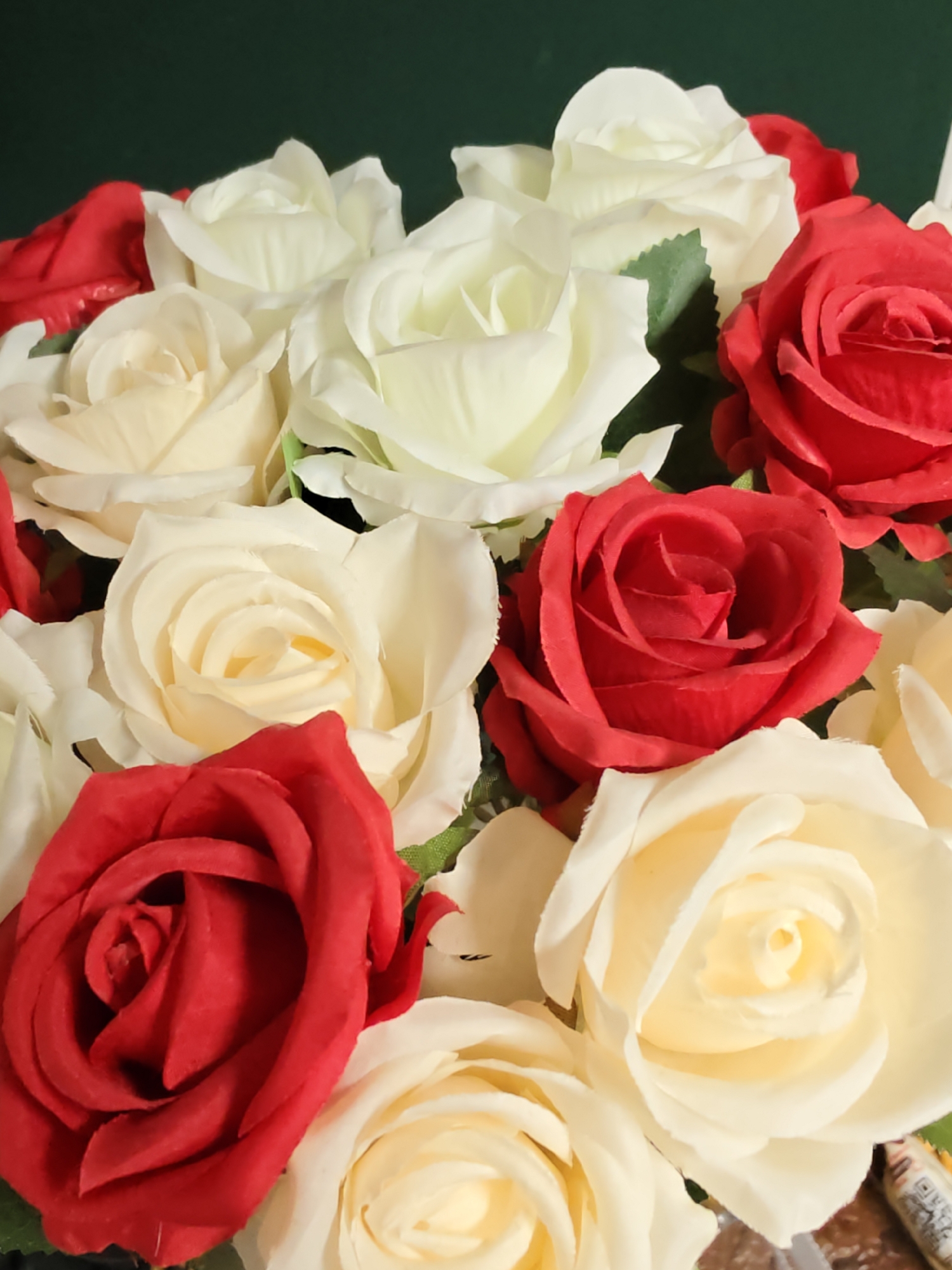 A close-up, high-angle shot shows a bouquet of red and white artificial roses against a dark green background. The roses are arranged in a dense cluster, filling the frame. The lighting is bright, with some shadows emphasizing the texture of the petals. There are no people or animals present, and the environment is unclear beyond the dark background. No specific location in Petah Tikva, Israel can be determined from the image.