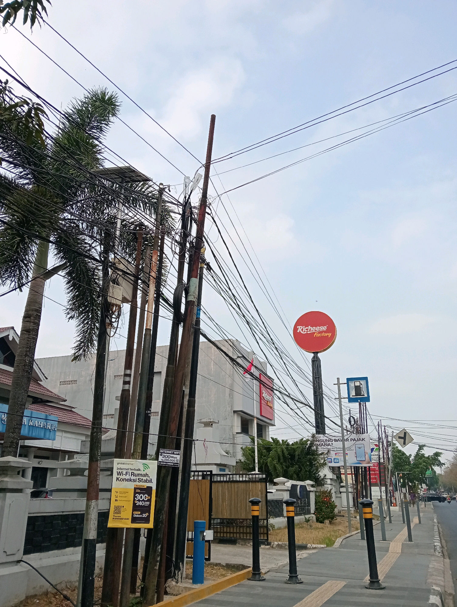 The image depicts a street scene in Samarinda, Indonesia, with a focus on a busy intersection of utility poles and overhead wires. Palm trees are visible on the left side of the image, with their fronds entangled in the dense network of wires. A large red circular sign with the "Richeese Factory" logo is prominently displayed on a pole. A blue sign with a gas pump icon is also visible. The street is paved, with a sidewalk featuring tactile paving for the visually impaired. Several utility poles, some made of wood and others of metal, are clustered together, supporting a multitude of electrical and telecommunication wires. A building with a grey facade is visible in the background, and a few cars are driving on the road. A yellow advertisement poster for "globalxtreme" Wi-Fi is attached to one of the poles, offering "Wi-Fi Rumah, Koneksi Stabil." The sky is overcast with scattered clouds.