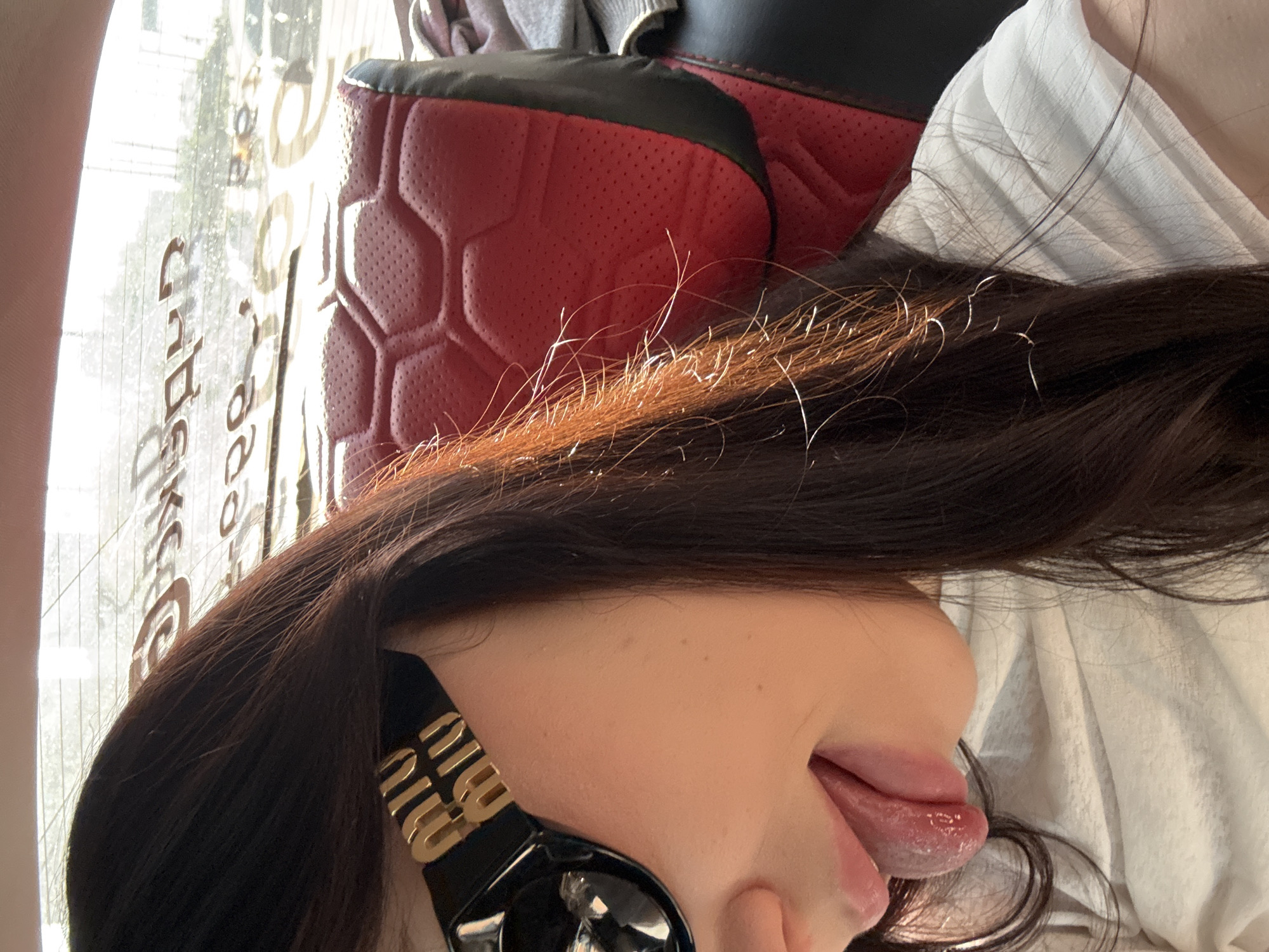 A young woman is leaning towards the camera inside a car. She is wearing large, black sunglasses with gold Miu Miu logos on the sides. Her tongue is sticking out. Her dark hair is long and hangs over her shoulders. The background shows the red quilted leather headrest of a car seat, and through the rear window, a sunlit urban landscape is visible, with trees and buildings. The image appears to have been taken in Krasnogorsk, Russia, based on the context provided.