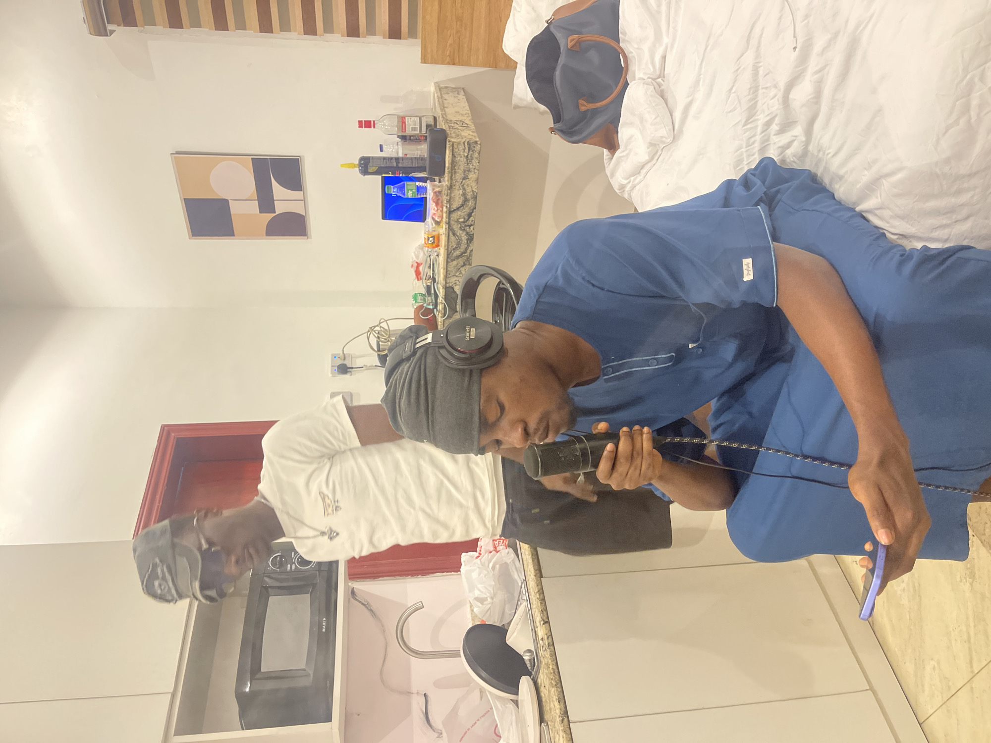 Two men are in a room that appears to be a hotel room or a domestic setting with a bed. One man, wearing a blue outfit and headphones, is seated and holding a microphone to his mouth, as if speaking or recording. The other man, wearing a white t-shirt and sunglasses, stands behind him.
On a counter behind them, there are several bottles, a laptop displaying a blue screen, and some plastic bags. A microwave is visible below the counter. A piece of abstract art hangs on the wall. The lighting suggests it is daytime. The room is clean and organized. The overall mood seems casual and focused on the recording activity.