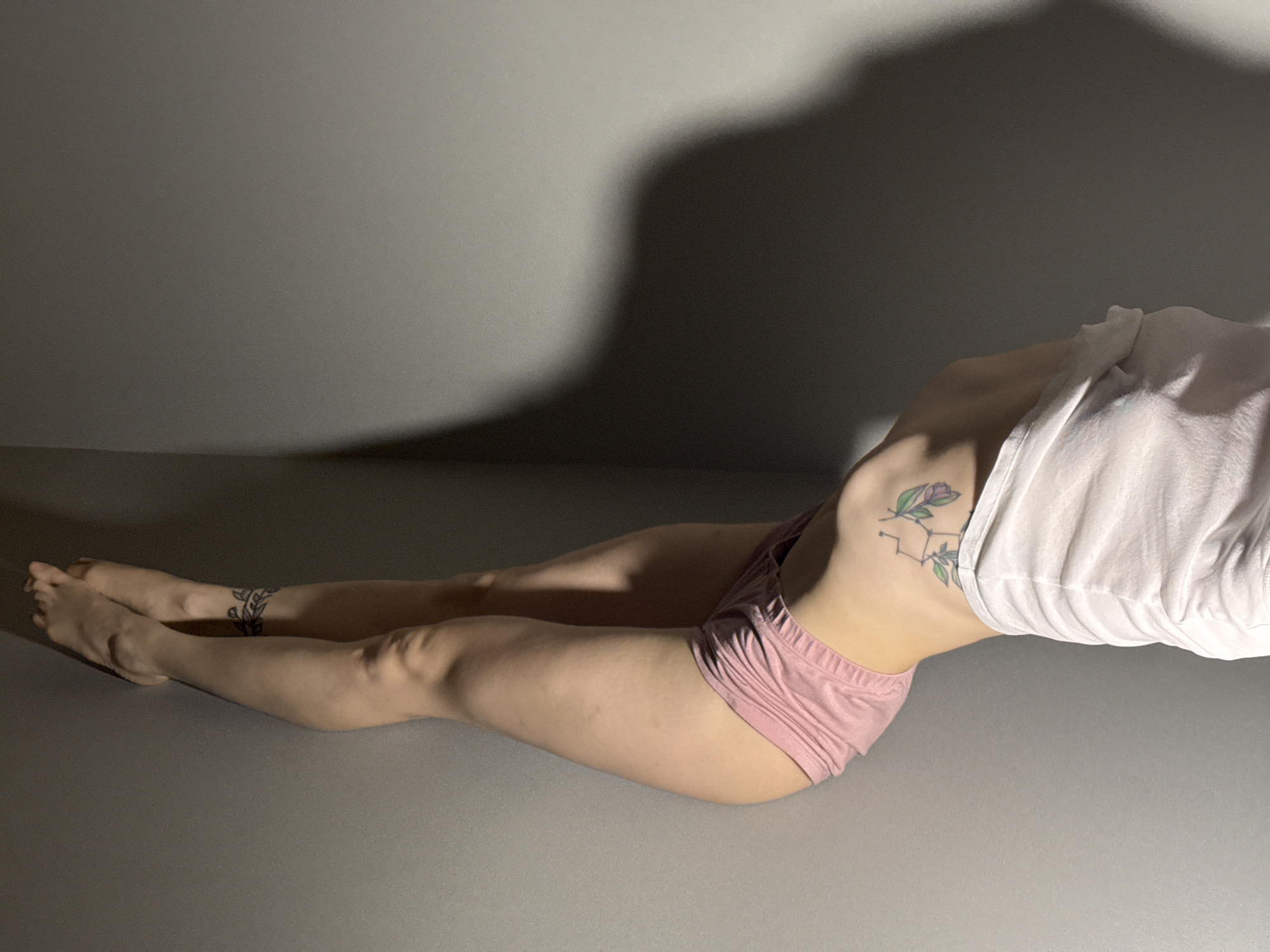 A person is captured in a reclining or stretching pose, likely indoors against a minimalist backdrop. The subject, who appears to be female, is positioned on a plain grey surface, possibly a floor or mat, with a matching grey wall behind. She is wearing light pink underwear or shorts and a loose white t-shirt that is partially lifted, exposing her midriff. Her body is arched in a graceful curve, suggesting flexibility or a state of relaxation.
Notable details include two tattoos: one on her left ribcage, depicting a floral or plant-like design, possibly integrated with a constellation or geometric pattern, and another band-like tattoo with leafy motifs on her lower left leg or ankle. Her legs are extended outwards, and her feet are relaxed.
Strong, dramatic shadows are cast on the wall and floor, emanating from the person's upper body and head, indicating a single, focused light source positioned to the right of the frame. This lighting creates a high-contrast aesthetic and depth. The setting provides no specific clues regarding the time of day or weather, as it appears to be an interior space with artificial or controlled lighting. No text is visible within the image. The provided context indicates the location is Chaikovsky, Russia, although this is not visually identifiable in the scene.