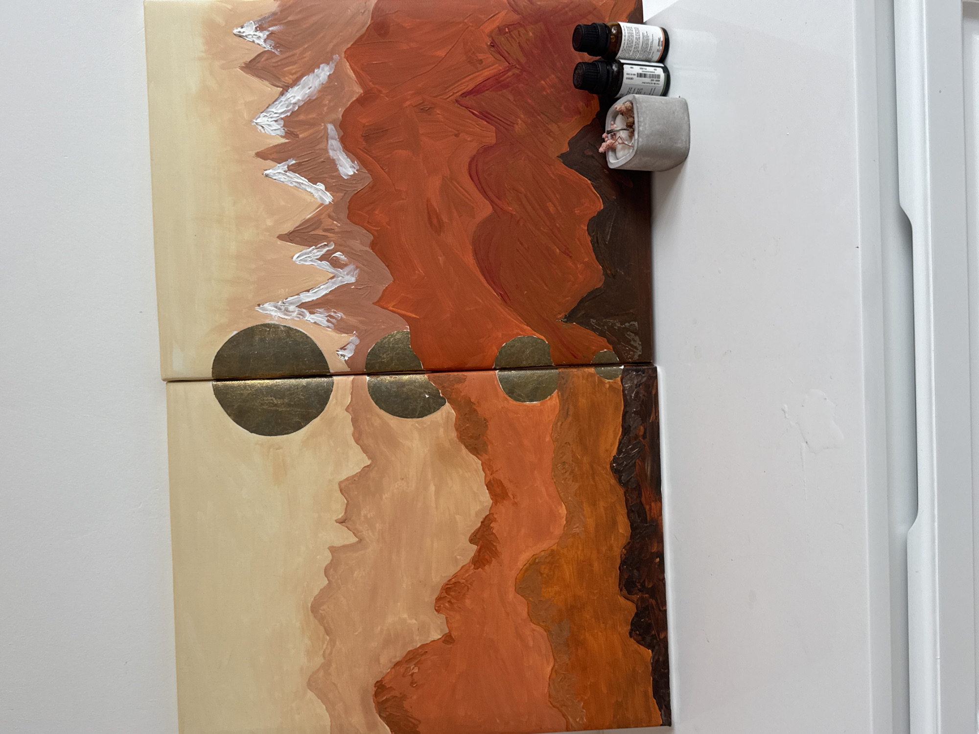 The image shows two painted canvases attached to the side of a white refrigerator. The paintings appear to be an abstract landscape in earth tones (tan, brown, rust). The upper painting has white paint strokes. The two paintings are placed in alignment to give the impression of one larger painting, with gold circles painted across the divide. Three small bottles and a small concrete pot holding dried flowers are magnetically attached on the refrigerator next to the artwork. The location is unknown and there is no indication of Bac Nha Trang Ward, Vietnam.
