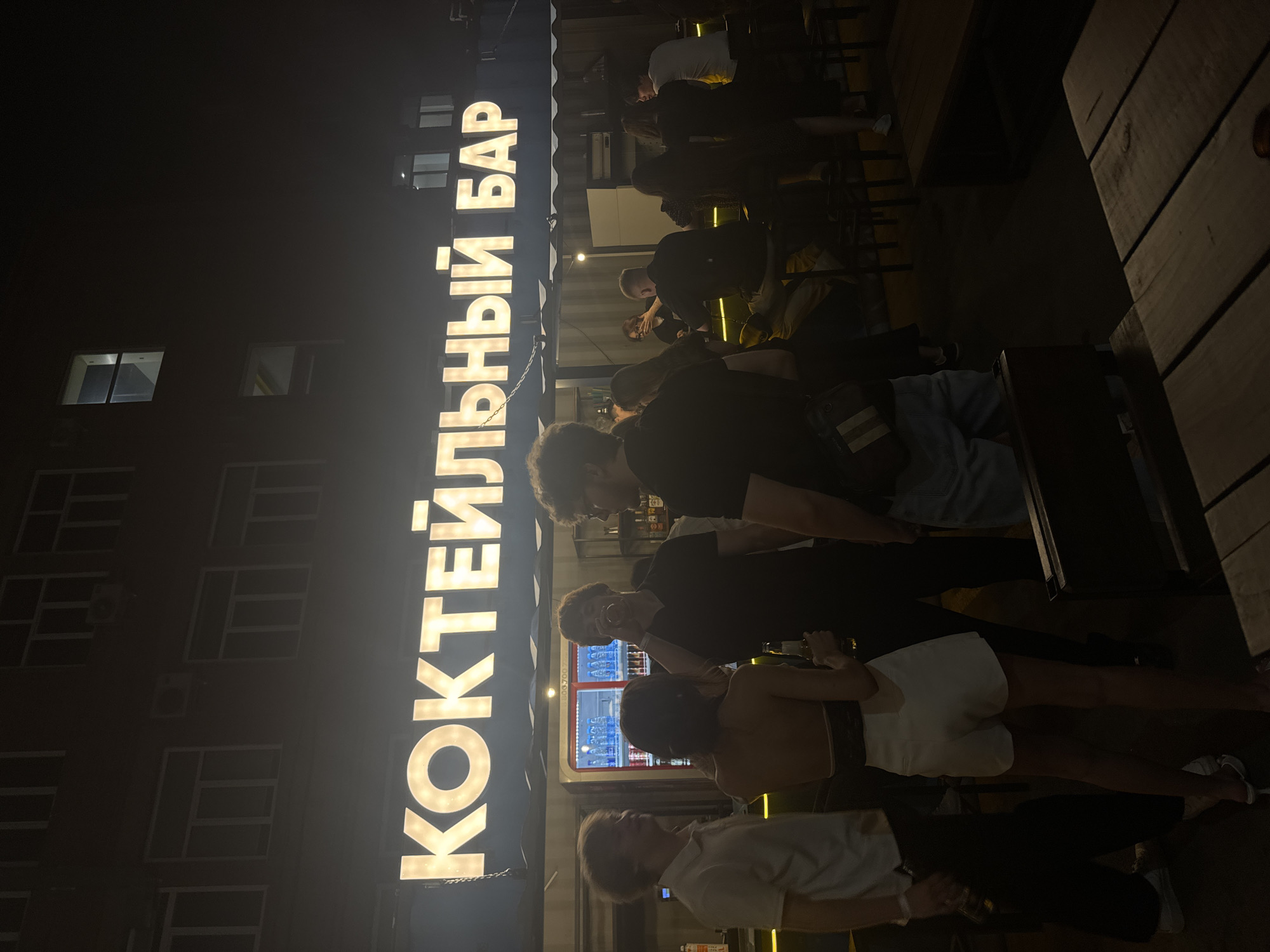 The image depicts an outdoor bar scene at night in Kemerovo, Russia. A prominent, illuminated sign displaying the Cyrillic text "КОКТЕЙЛЬНЫЙ БАР" (Cocktail Bar) is mounted above the bar structure, which appears to be constructed from dark, modular units or shipping containers.
Approximately 10-15 individuals are present within the scene, engaged in social interaction. In the midground, a group of four individuals stands in front of the bar. From left to right: a man with light-colored hair, wearing a white polo shirt, stands facing left; a woman with dark hair, wearing a white strapless top and white shorts, is seen from behind, holding a bottle in her right hand; a man with dark, curly hair and a beard, wearing a black t-shirt, is actively drinking from a clear glass; and a fourth man, also in a dark t-shirt and wearing glasses, is partially visible on the far right of this group. Behind them, inside the bar, a commercial refrigerator stocked with bottled beverages is visible, along with a partially obscured phone number "8.900.700" on an adjacent surface.
Further to the right, several other patrons are seated on stools at an illuminated bar counter, facing a presumed bartender or bar staff member. Bottles of alcohol are visible on shelves behind the counter.
The background features a dark, multi-story building with numerous rectangular windows, some of which exhibit faint internal illumination. An air conditioning unit is mounted on the building's exterior. The foreground shows sections of dark wooden tables and benches, indicating an outdoor seating area. The overall scene is dimly lit, with the bar sign providing the primary source of illumination.