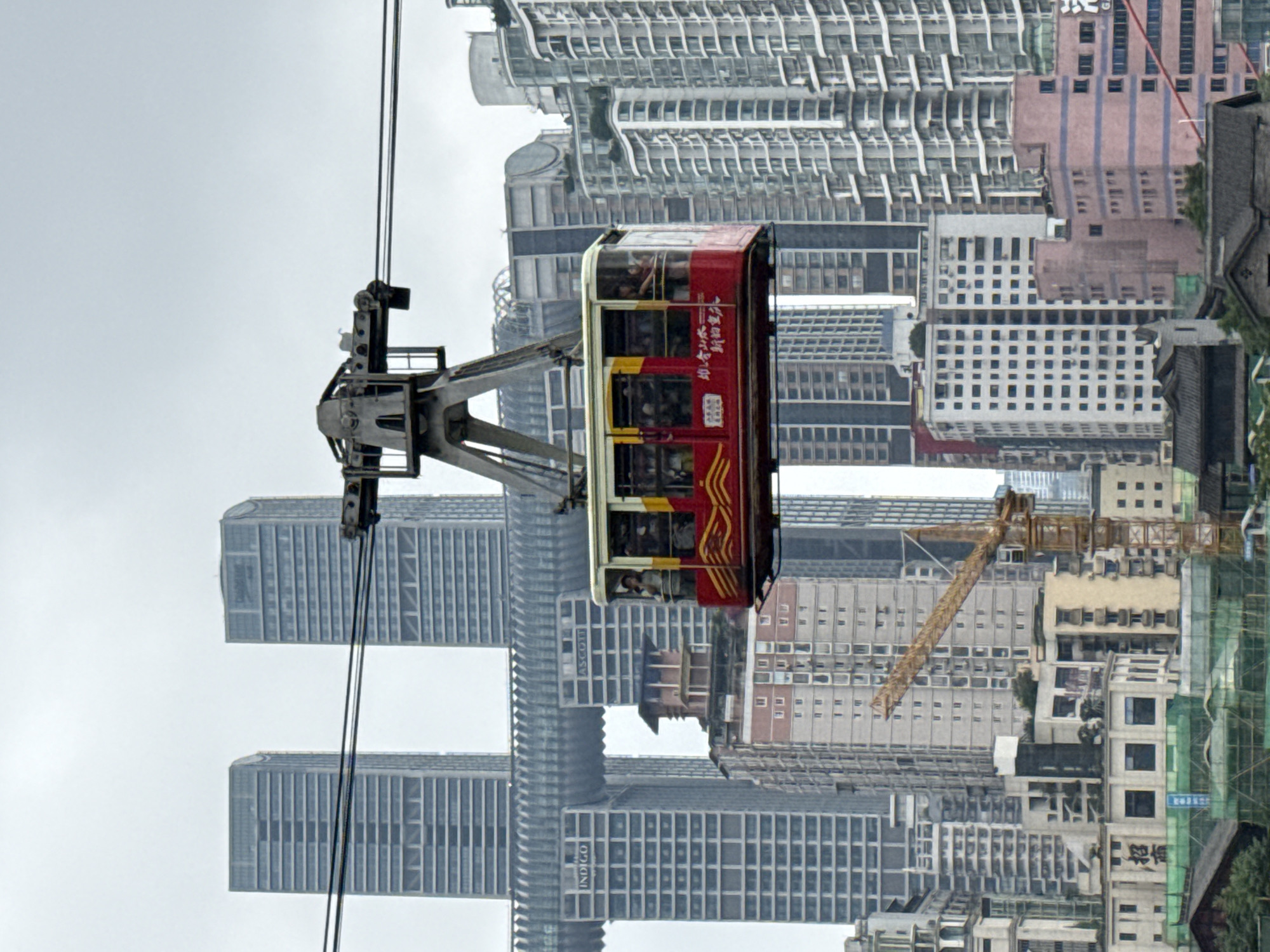 The image depicts a cable car suspended in front of a cityscape in Yuzhong District, China. The cable car is red and white with visible occupants inside. The cityscape is characterized by numerous high-rise buildings, some with recognizable architectural features and varying colors. A construction crane is visible among the buildings. The sky is overcast. The image is rotated clockwise 90 degrees.
