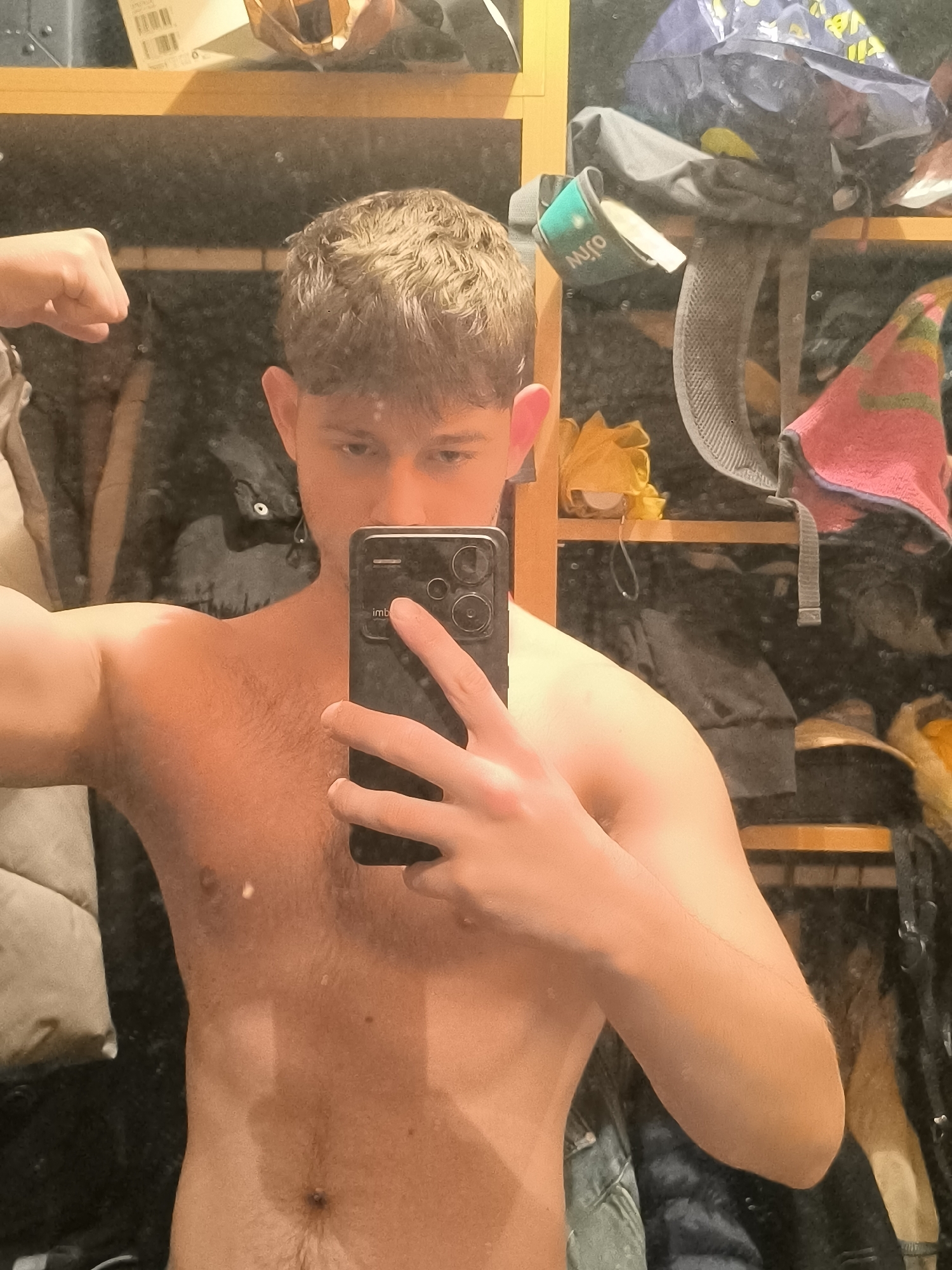 A shirtless young man with brown hair is taking a selfie in front of a mirror. He is holding a black smartphone with multiple camera lenses. He is flexing his left arm. The background shows a cluttered shelf with various items, including bags, clothing, and other objects. There is no information in the image to indicate that this photo was taken in Sofia, Bulgaria.