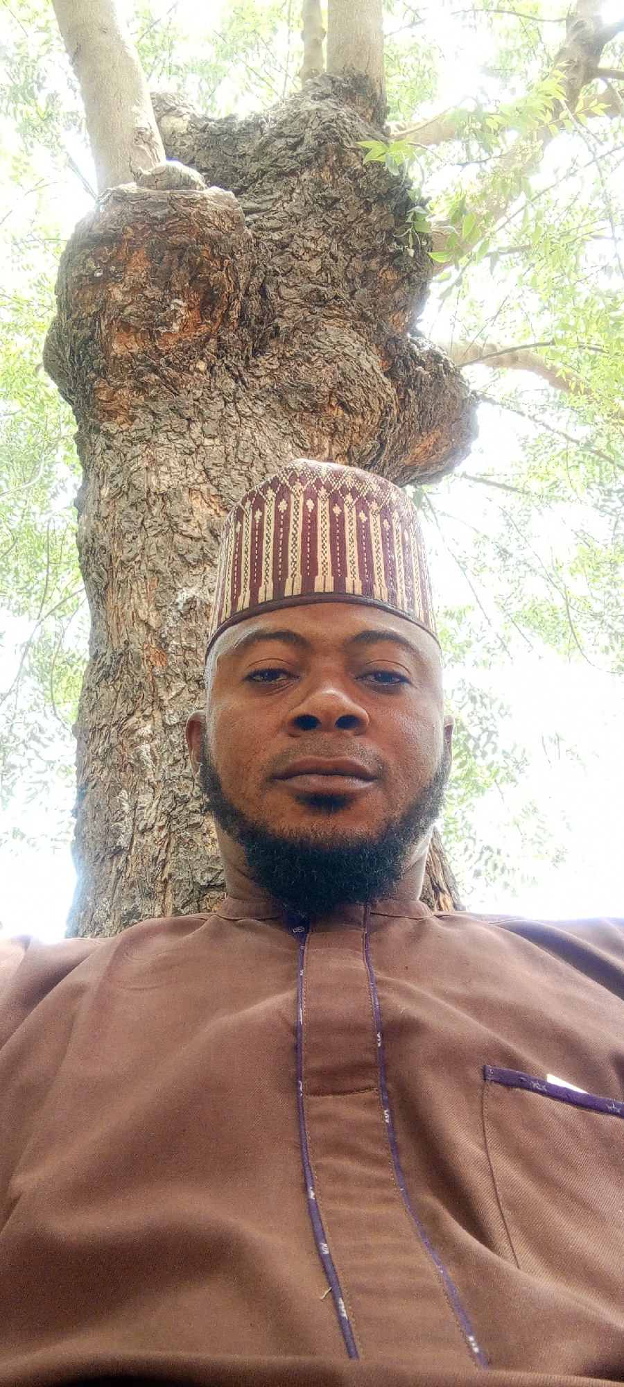A close-up shot features a man wearing a brown traditional outfit and a patterned cap. He has a dark beard and appears to be outdoors, with a large tree trunk visible behind him. The environment suggests a natural setting, with green leaves and foliage visible in the background, indicative of an outdoor space in Gawuna, Nigeria.