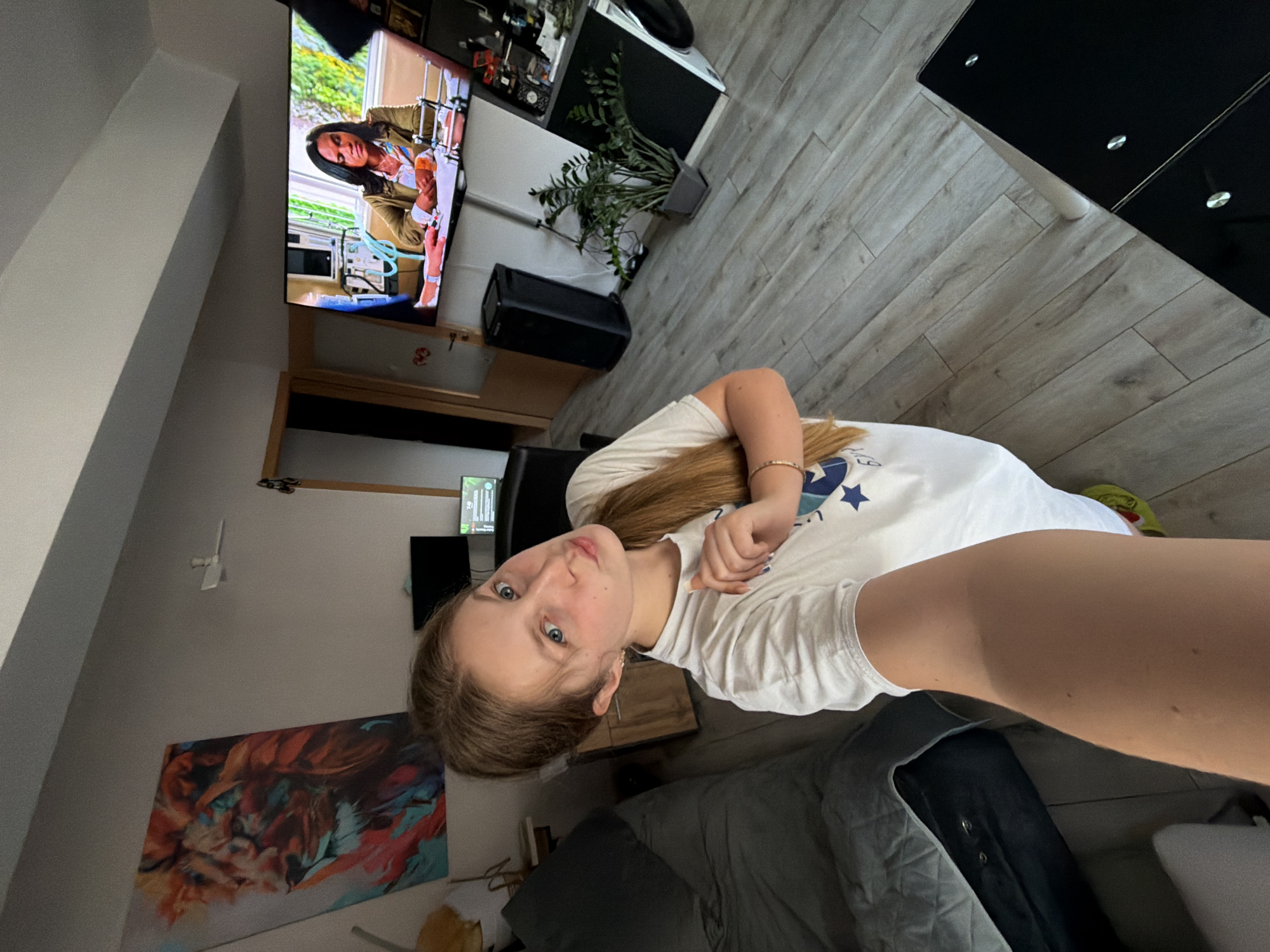 A young woman is captured in a selfie taken indoors, appearing to be in a living room or bedroom. The image is rotated counter-clockwise, showing the subject with her head tilted towards the camera, her light brown hair pulled back, and striking blue eyes. She wears a white t-shirt adorned with a visible blue star logo on her right shoulder and chest area. A slender bracelet is visible on her left wrist. Her expression is neutral and direct.
In the background, behind her to the left, a wall features a large, colorful abstract painting with shades of orange, teal, and brown. A white wall-mounted device is also visible above the painting. To her right, a modern flat-screen television is mounted on the wall, displaying a scene with a dark-haired woman in a beige or olive jacket, holding an orange object and interacting with what appears to be medical equipment, including tubing and machines. Beneath the TV, a wooden shelf or cabinet is visible.
The room features light-colored laminate flooring and a dark, possibly black, glossy table or cabinet with silver accents. A potted green plant sits on a surface near the television, and a black speaker is positioned on the floor. Part of a dark grey quilted blanket or cover is visible in the lower left corner of the frame. The overall setting suggests a modern home interior in Burgas, Bulgaria. No specific time of day or weather can be discerned from the indoor scene.