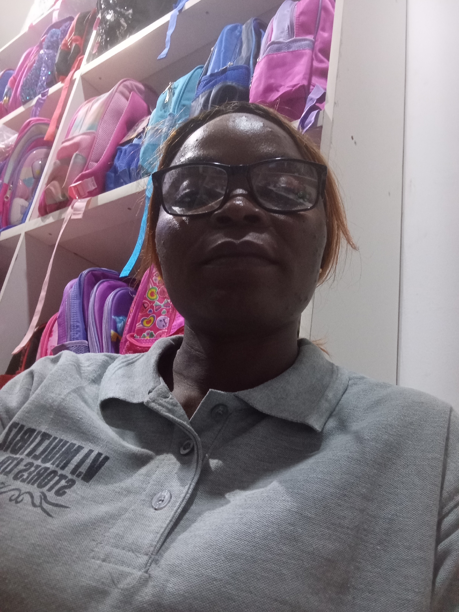 In Howayi, Nigeria, a woman with dark skin and short, reddish-brown hair wears glasses and a grey collared shirt with a printed logo. She is captured in a close-up, self-taken photograph with a shallow depth of field. The background is cluttered with brightly colored backpacks arranged on shelves, suggesting a retail or storage environment for school supplies. The lighting appears to be artificial, casting shadows and highlighting the textures of the woman's clothing and skin. The overall impression is that of a casual, personal snapshot taken within a commercial setting.