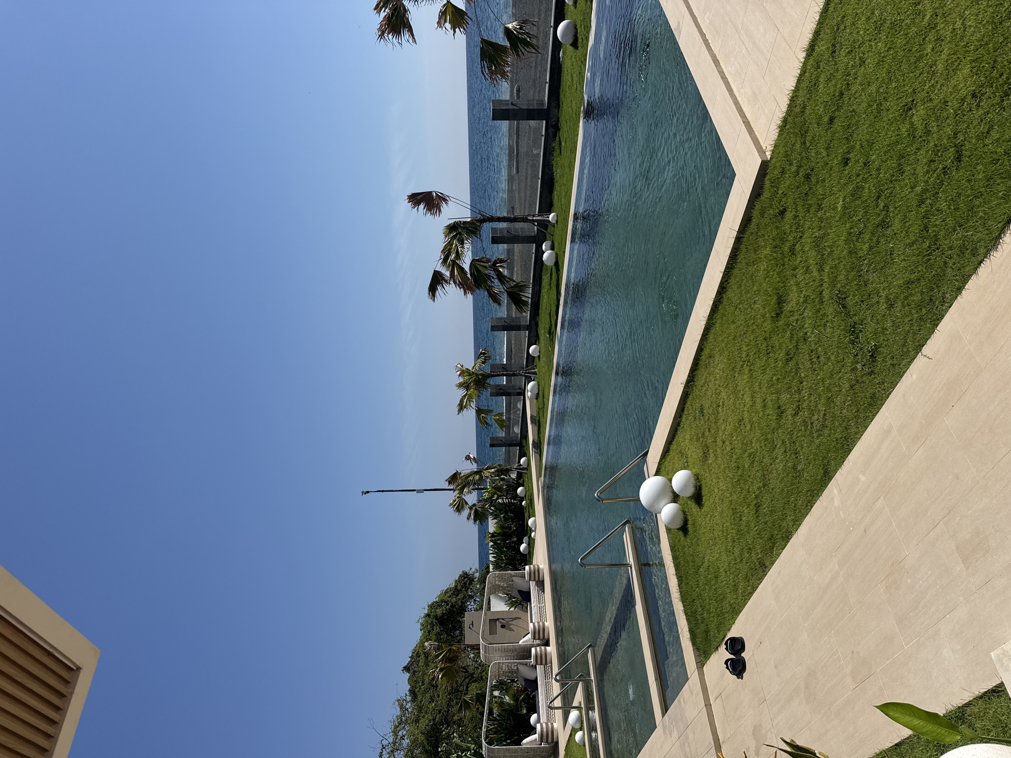This is a full, eye-level, outdoor shot of a modern, resort-style swimming pool area next to the ocean. The scene is set on a clear, sunny day with a vibrant blue sky and calm, dark blue ocean water. In the foreground, there is a light-colored tiled pathway leading to a well-maintained green lawn. On the pathway, a pair of black flip-flops are placed neatly. Adjacent to the pathway is a swimming pool with turquoise water and several white spherical decorative objects scattered around its edge.
The pool is a geometric shape with a shallow section and a deeper main area, featuring chrome ladder handles for access. Beyond the lawn is a black sand beach, and further out, the ocean stretches to the horizon. A low, modern fence made of dark stone pillars and glass panels separates the pool area from the beach. Several palm trees with fronds blowing in the wind are positioned along the beachfront, adding a tropical feel. In the background, to the left, a portion of a thatched-roof structure is visible, suggesting a covered seating or lounging area. The overall impression is one of a serene and luxurious coastal retreat in the city of Kubutambahan, Indonesia.