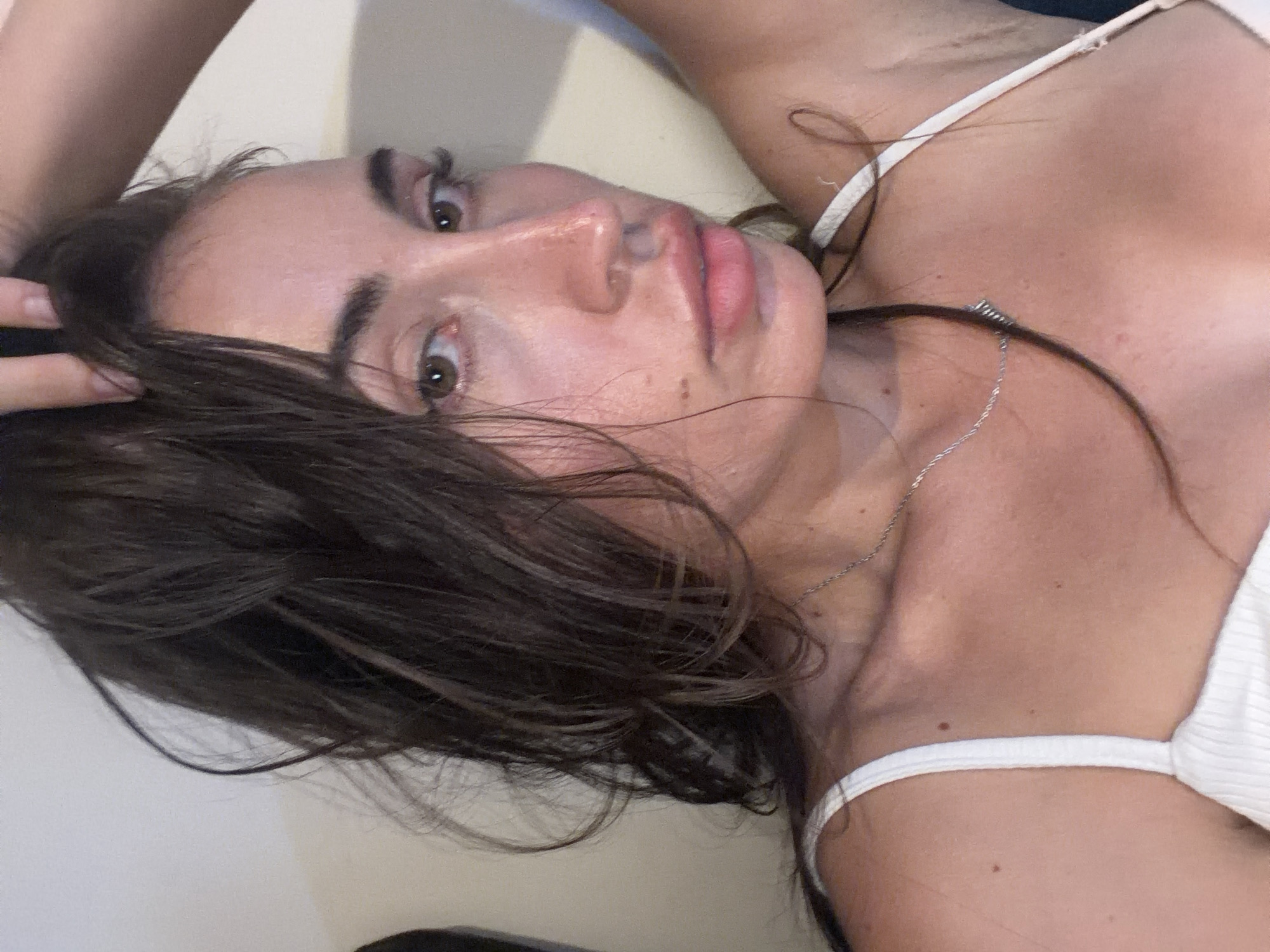 A medium close-up selfie shows a woman from the chest up. She has long, dark brown hair, which appears wet. Her eyes are light brown, and she is wearing a white ribbed tank top with thin straps. A thin silver chain necklace is visible around her neck. The background is a neutral, plain wall. The lighting suggests an indoor setting, possibly a home or a studio. The image is taken at a slight upward angle. The woman is located in Tersefanou, Cyprus.